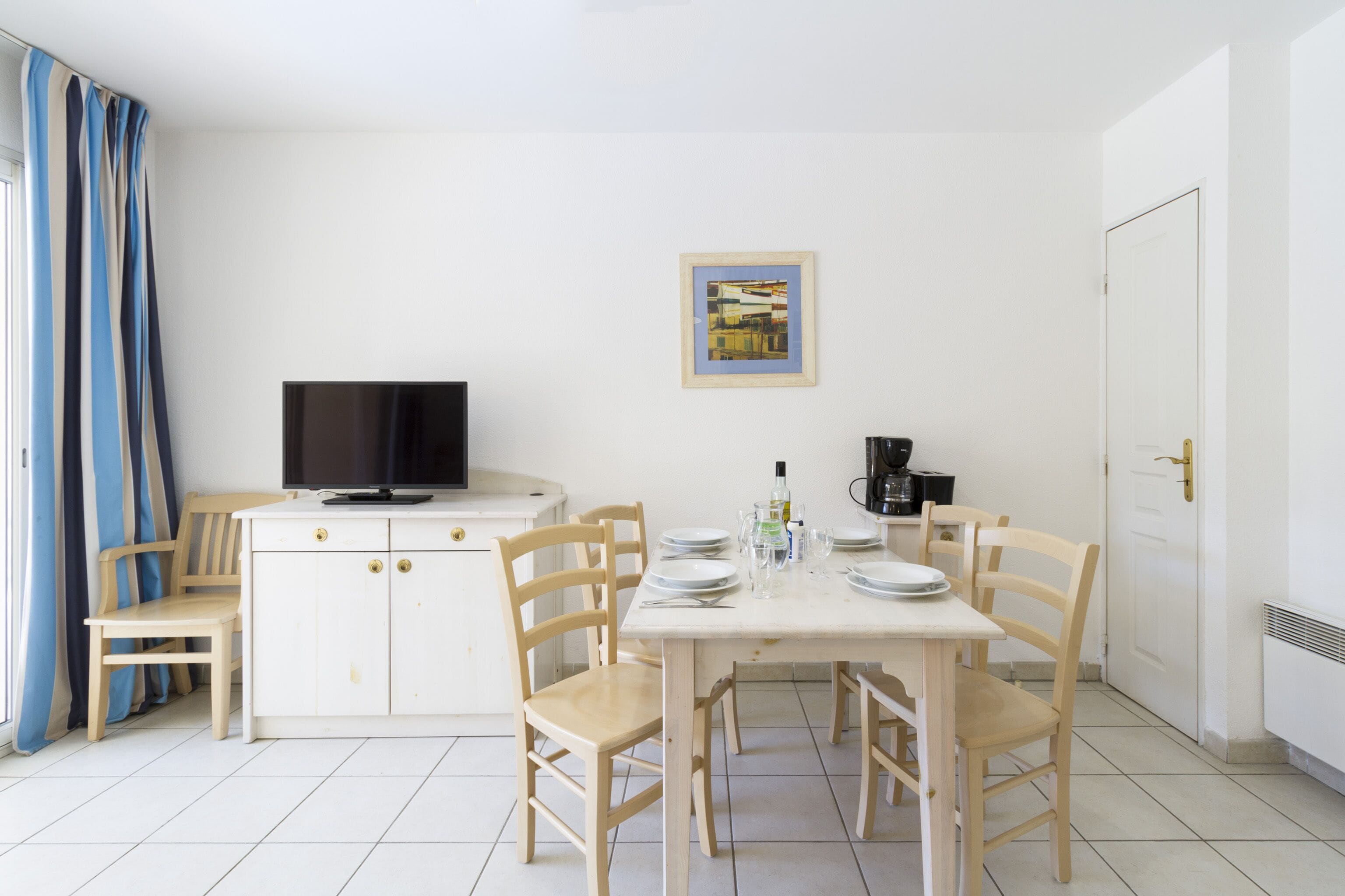 Een kleine, schone kamer met een eettafel voor vier personen, een televisie en een koffiezetapparaat.