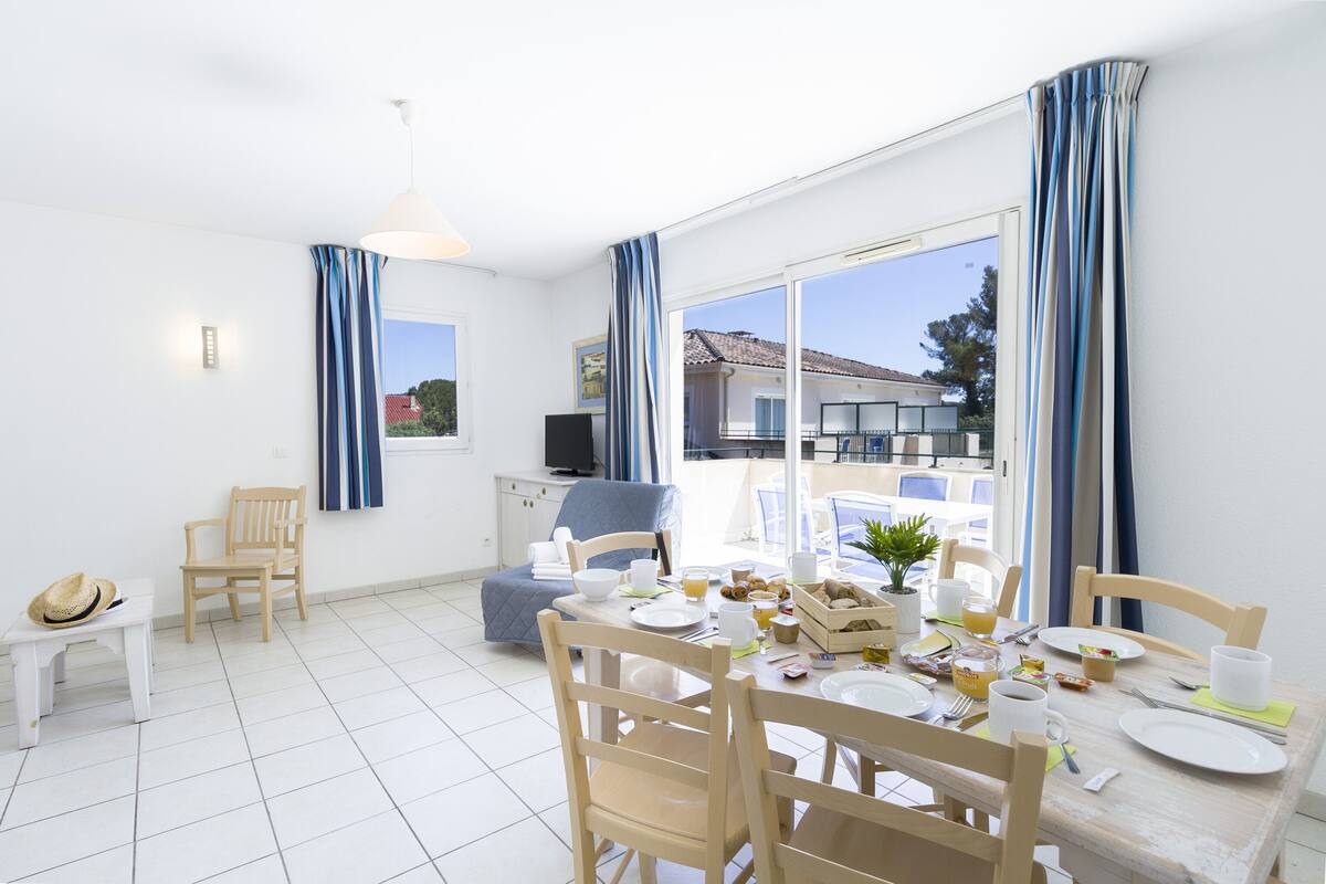 Un comedor con una mesa dispuesta para el desayuno, un televisor sobre un soporte y vistas al exterior.