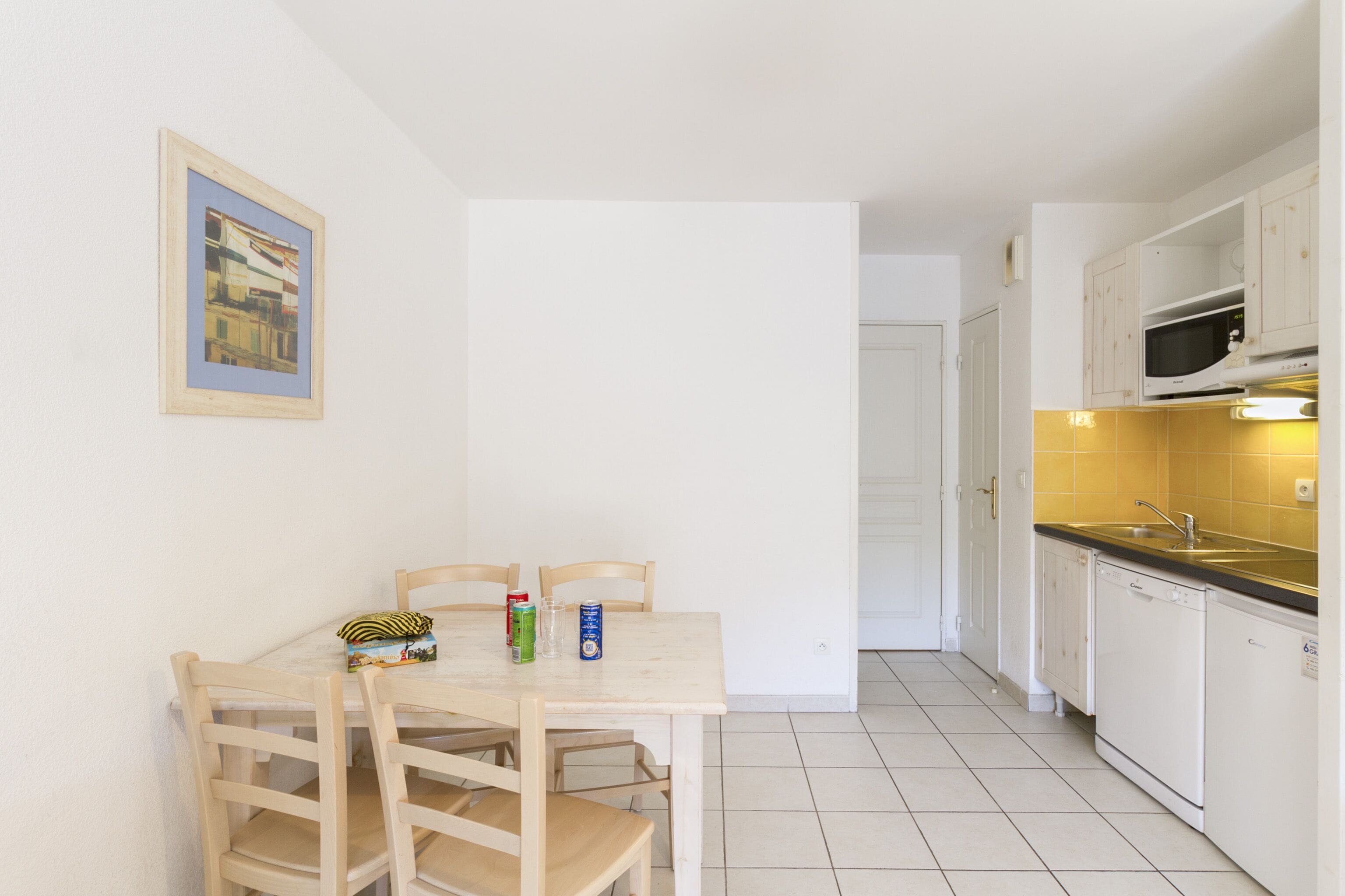 Eine kleine Küche mit weißen Schränken, einer Mikrowelle und einem Spülbecken. Es gibt einen Tisch mit Stühlen und ein gerahmtes Bild an der Wand.