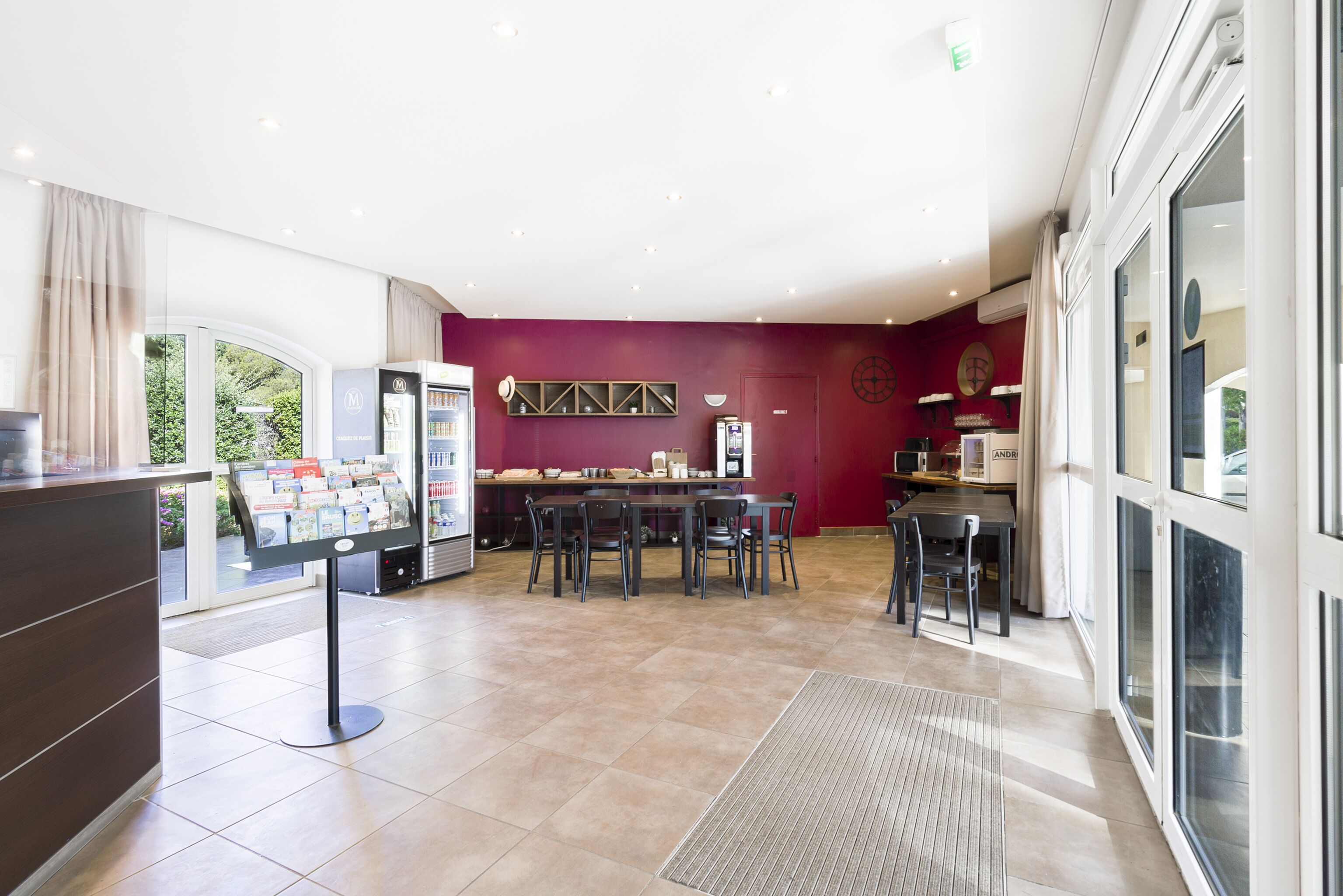 A modern hotel lobby with a reception desk, a self-service kiosk, a dining area with tables and chairs, and large windows offering a view of the outdoors.