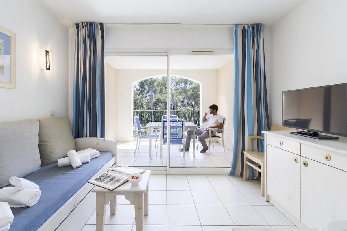 Habitación de hotel moderna con sofá, una mesa de comedor con un bol de fruta, televisión y balcón con vistas a árboles.