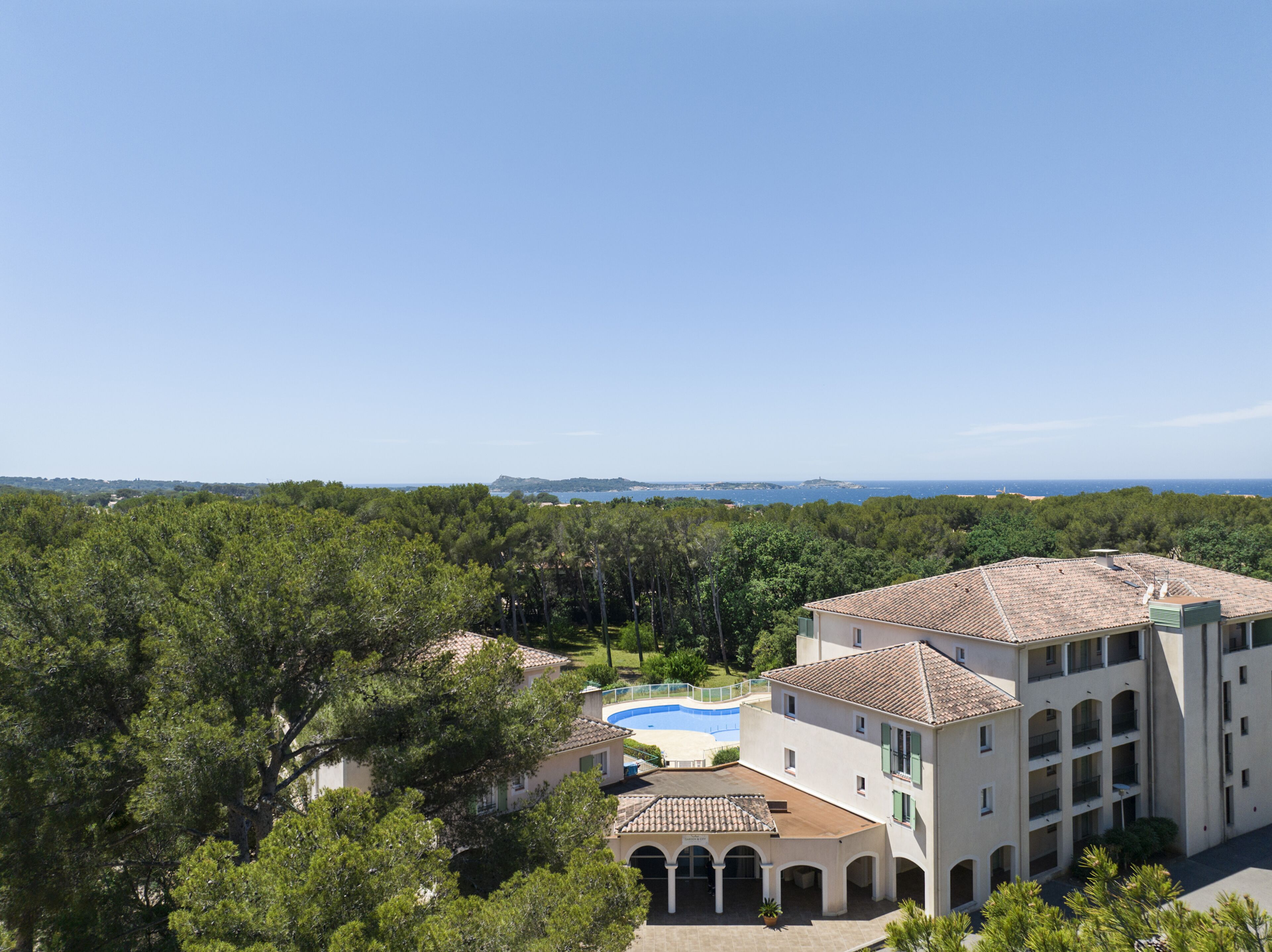 A hotel with a swimming pool, surrounded by lush greenery and a clear view of the sea.