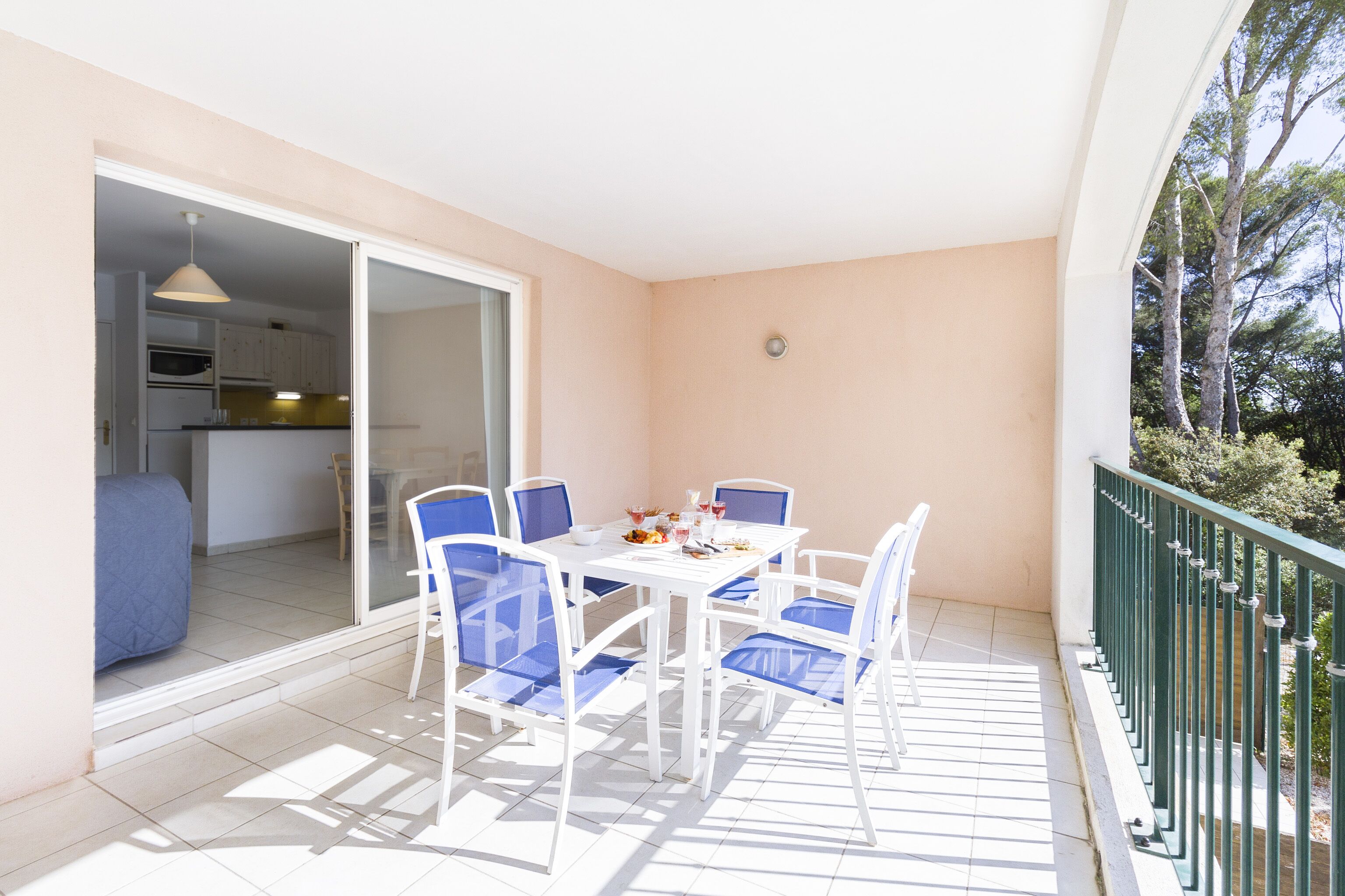 Un balcon avec une table blanche et des chaises bleues, une porte-fenêtre coulissante donnant sur une cuisine et une rambarde verte.