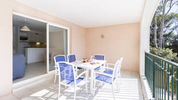 Un balcon avec une table blanche et des chaises bleues, une porte-fenêtre coulissante donnant sur une cuisine et une rambarde verte.