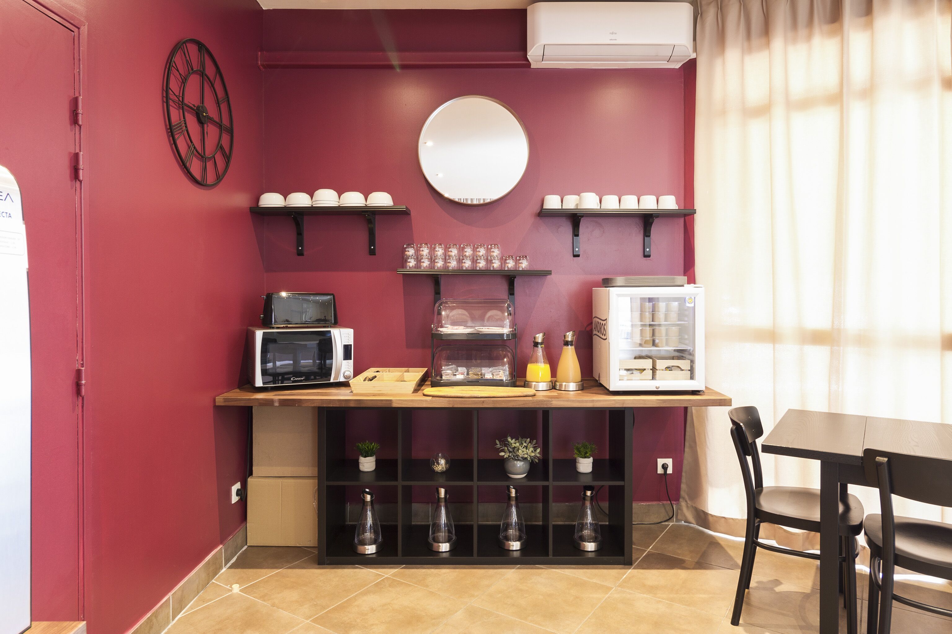 Une cuisine compacte équipée d’un four à micro-ondes, d’une machine à café et d’un réfrigérateur. On y trouve des étagères avec des tasses et une horloge au mur.