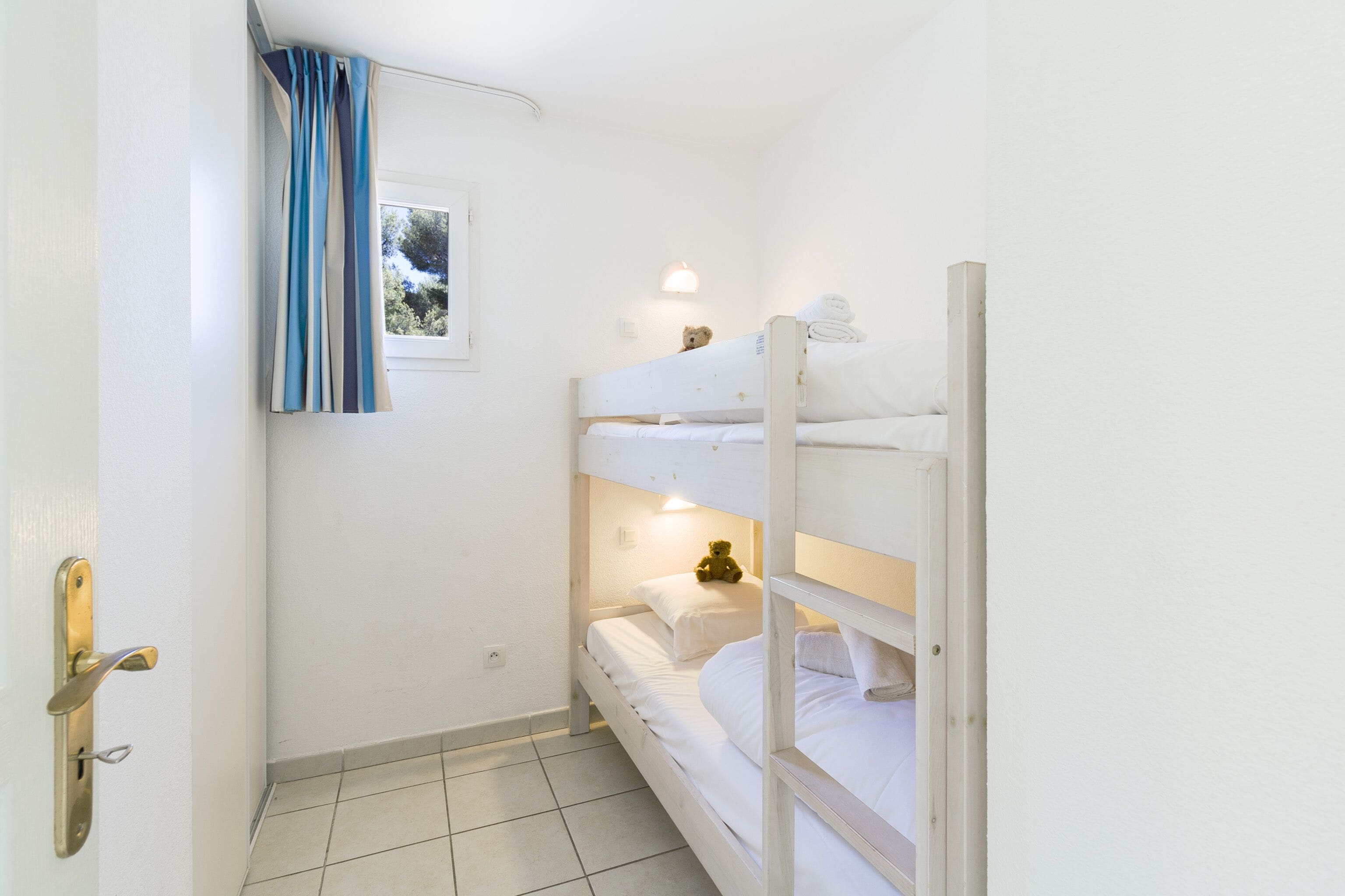 A small, clean room with two bunk beds, a window with curtains, a bedside lamp, and a teddy bear on the top bunk.
