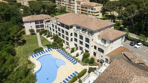 Un hotel de varios pisos con piscina rodeada de tumbonas, situado en una zona verde y exuberante, con vistas al mar y montañas a lo lejos.