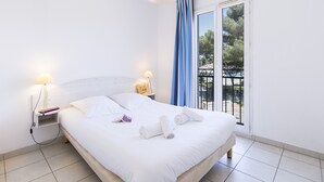 Una cama bien hecha con sábanas blancas y toallas enrolladas, una mesita de noche con lámpara, y vistas a un balcón con barandilla.
