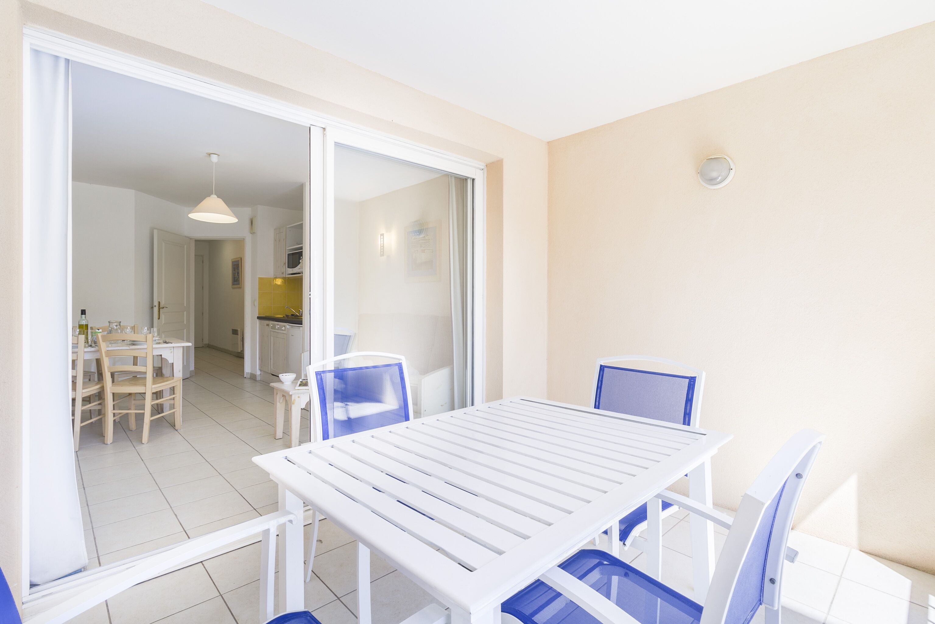 Un coin repas avec une table blanche et des chaises bleues, une porte-fenêtre coulissante donnant sur une cuisine et un luminaire au plafond.