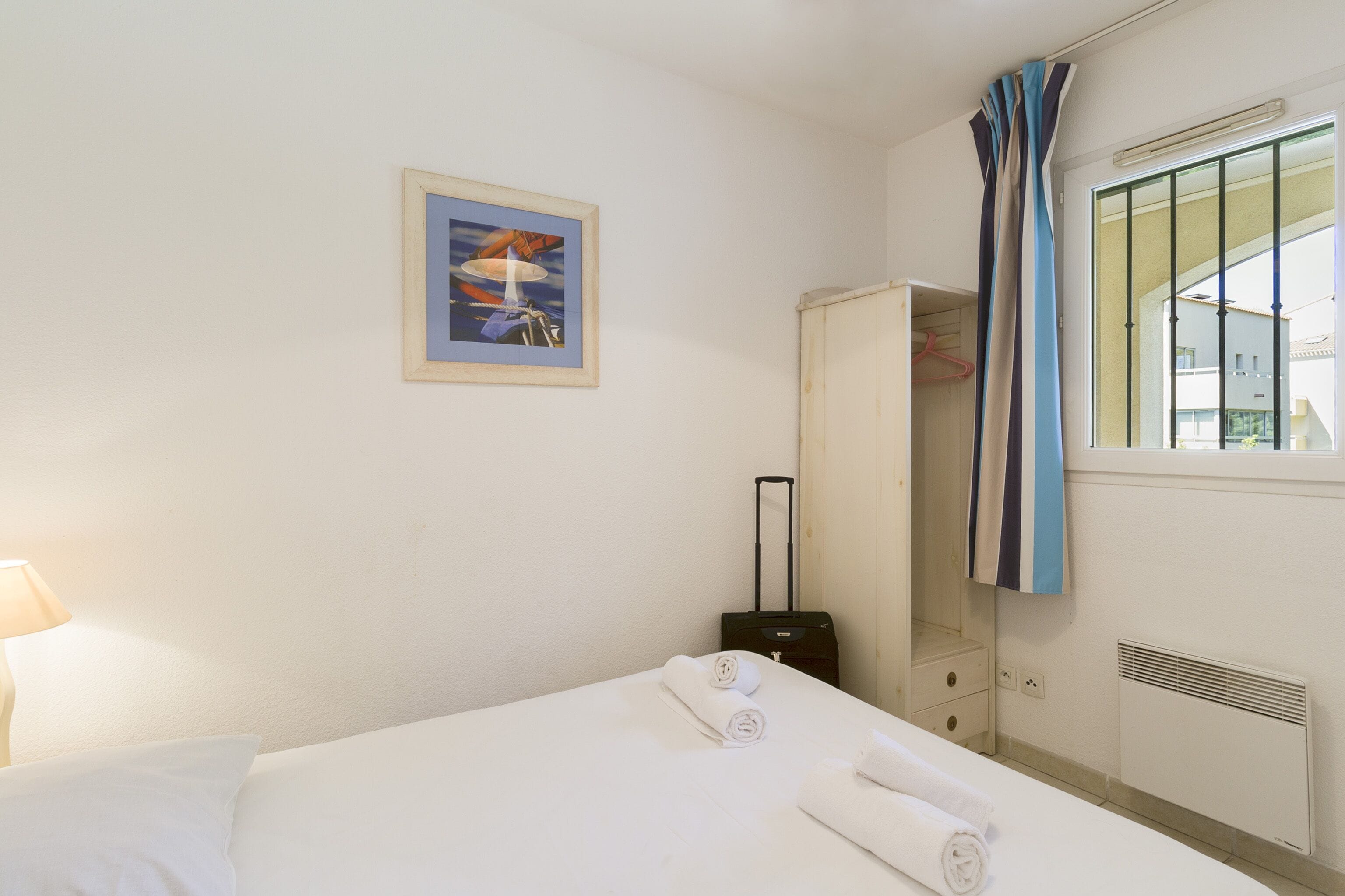 A hotel room with a bed, a suitcase, a wardrobe, a window with curtains, and a painting on the wall.