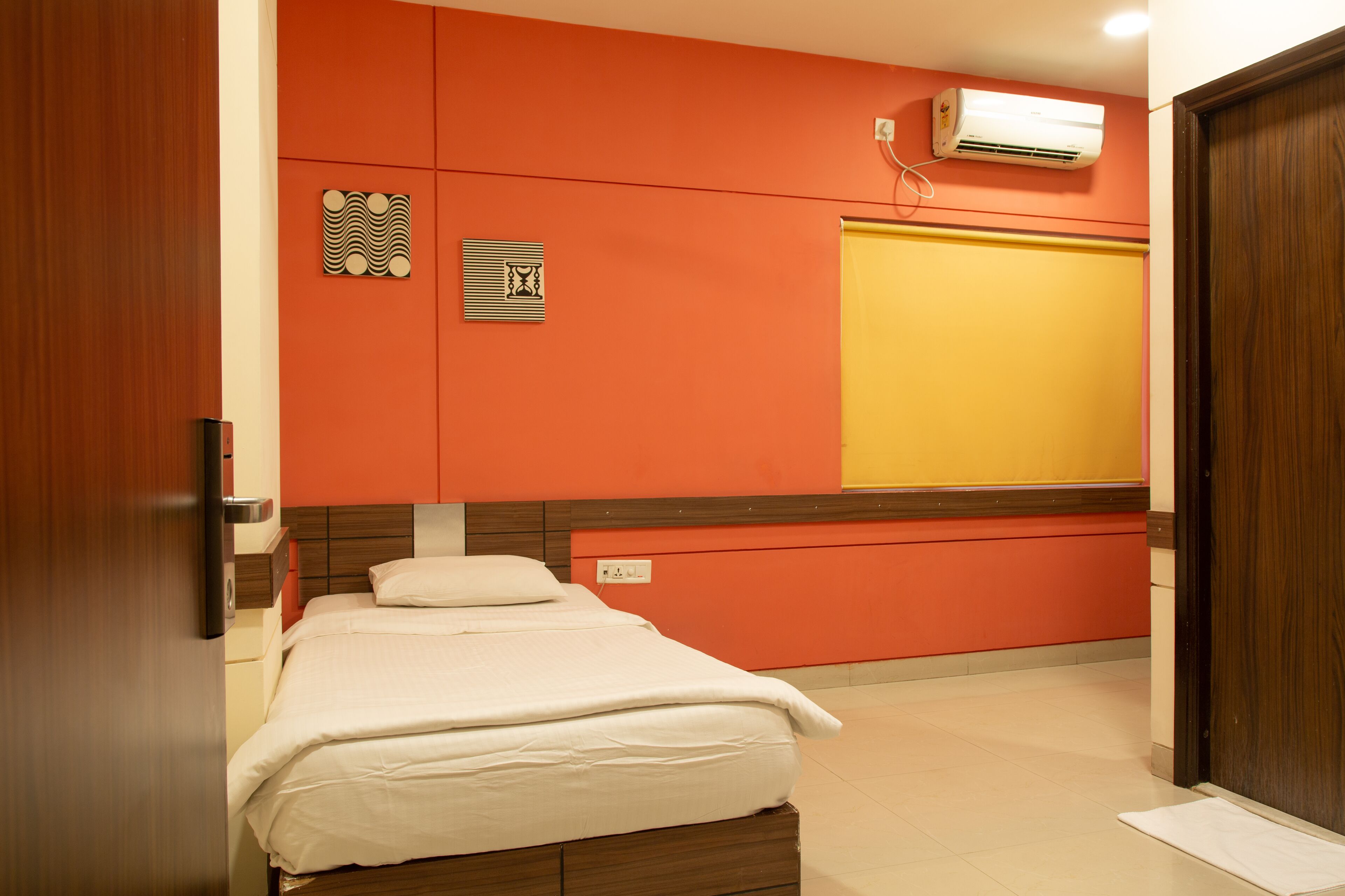 A hotel room with a bed, a wooden door, a wall-mounted air conditioner, and a yellow sliding door.