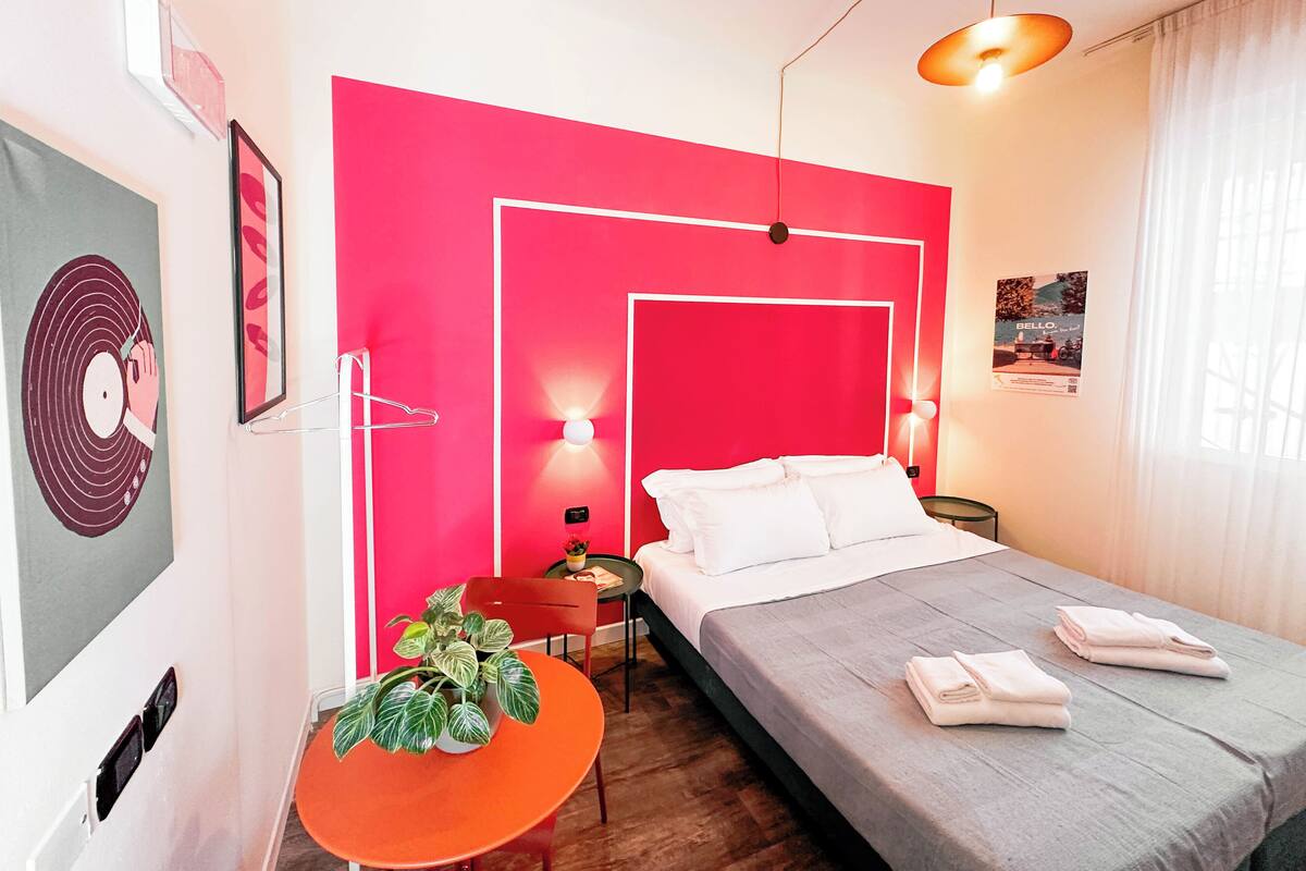 A modern bedroom with a bright red accent wall, a bed with white linens, a small orange table with a plant, and a nightstand with a lamp.