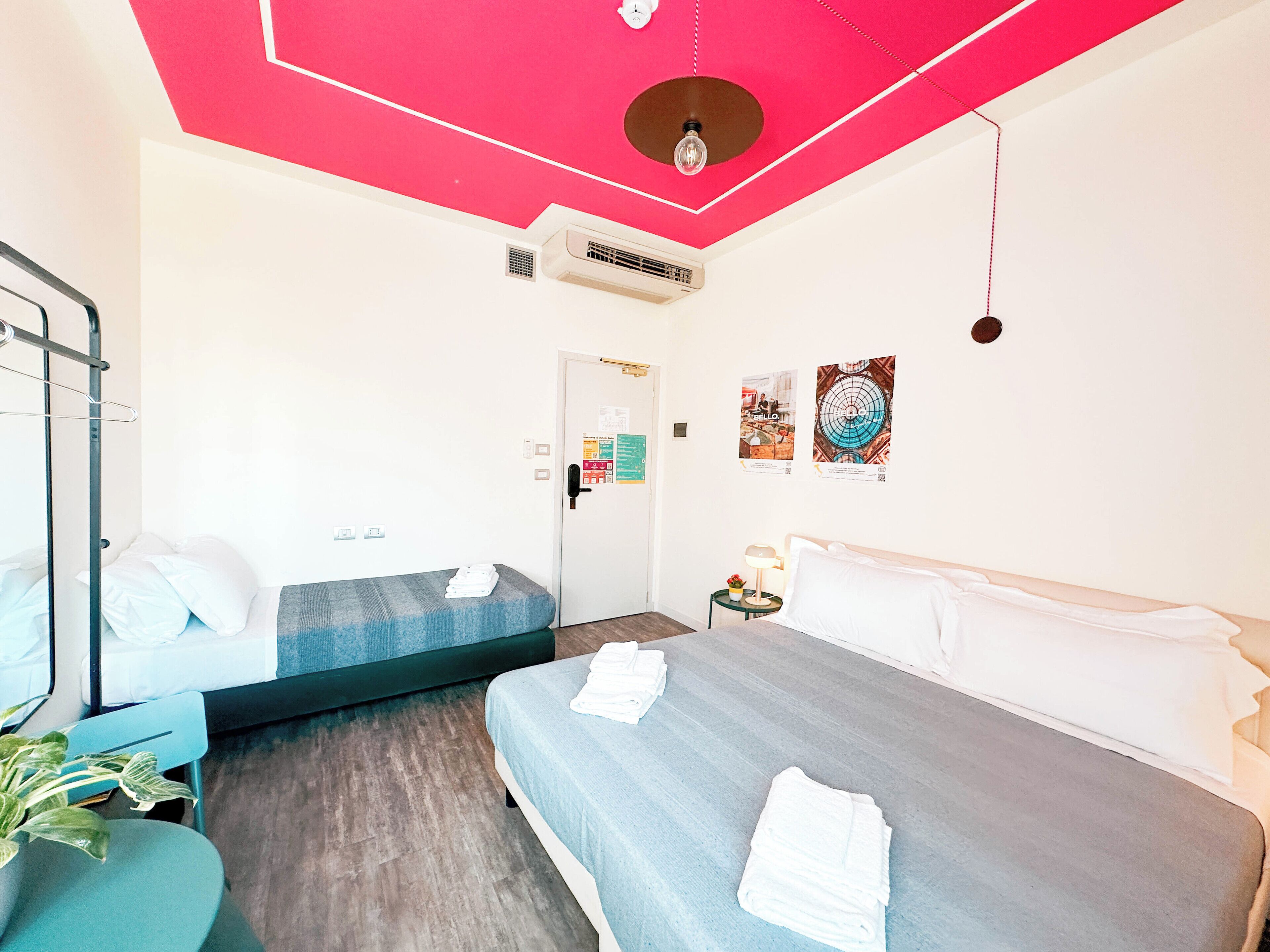 A room with two beds, a bunk bed, a ceiling fan, an air conditioning unit, a small table with a lamp, and a wall-mounted poster.