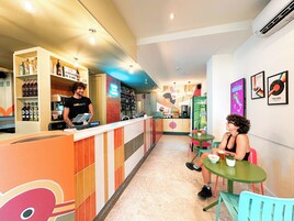 Uma área de bar com balcão, prateleiras com garrafas e uma pessoa atrás do balcão. Há cadeiras e mesas coloridas, e uma parede com quadros emoldurados.