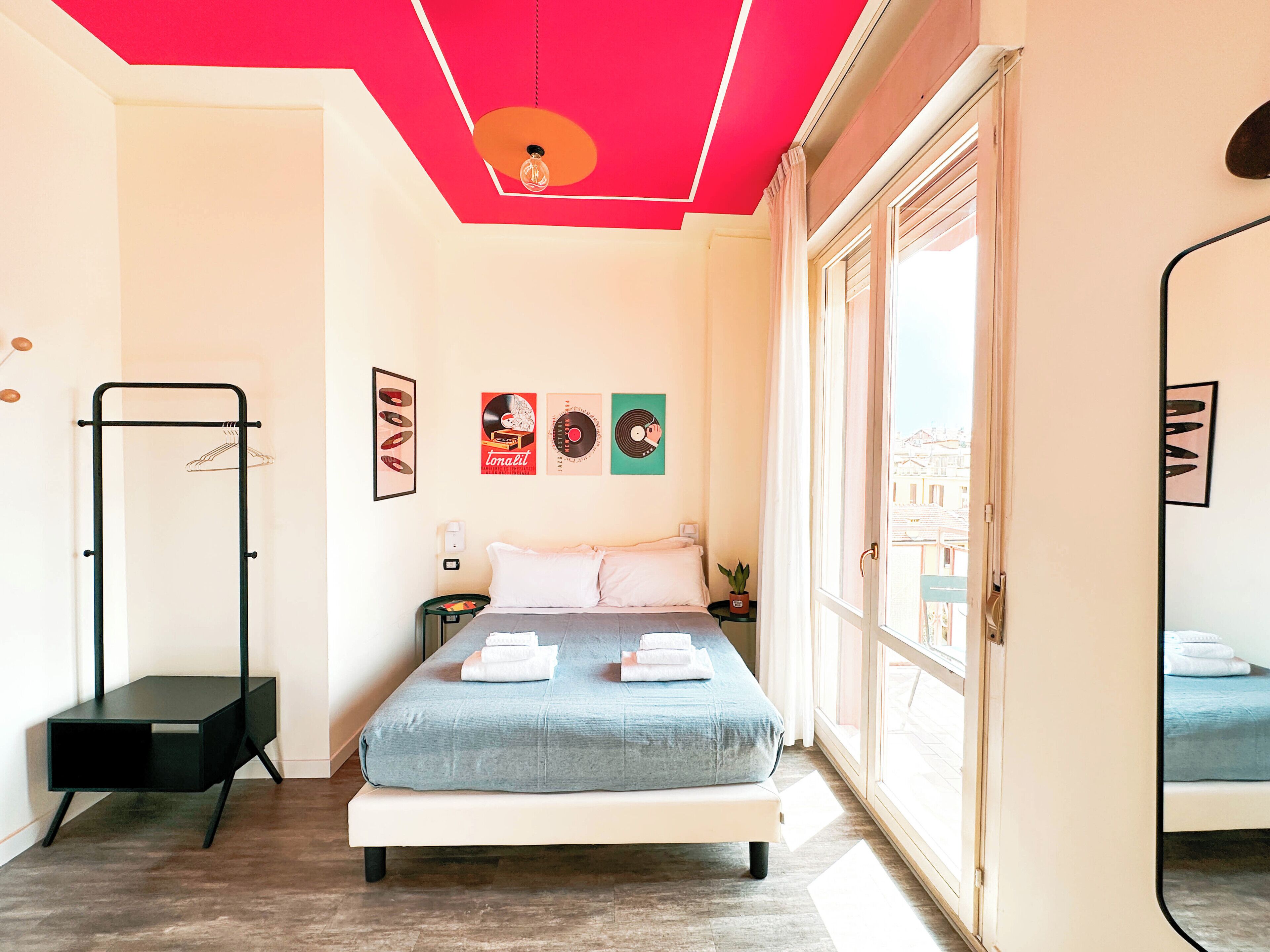 A bedroom with a bed, a bedside table, a hanging light fixture, a coat rack, and a window with a view.