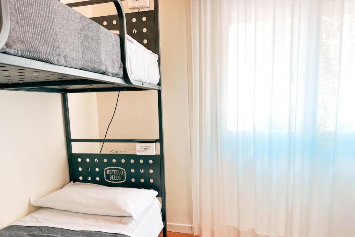 A bunk bed with a label "Ostello Bello" and a window with sheer curtains.