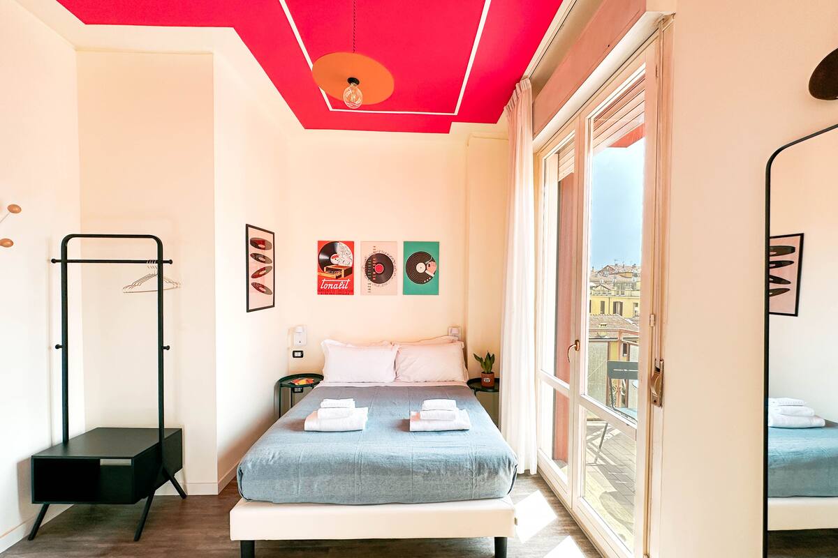 A bedroom with a bed, a bedside table, a hanging light fixture, and a window with a view of buildings.