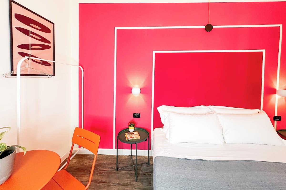 A modern bedroom with a large bed, a bright red accent wall, a small table with a plant, and an orange chair.