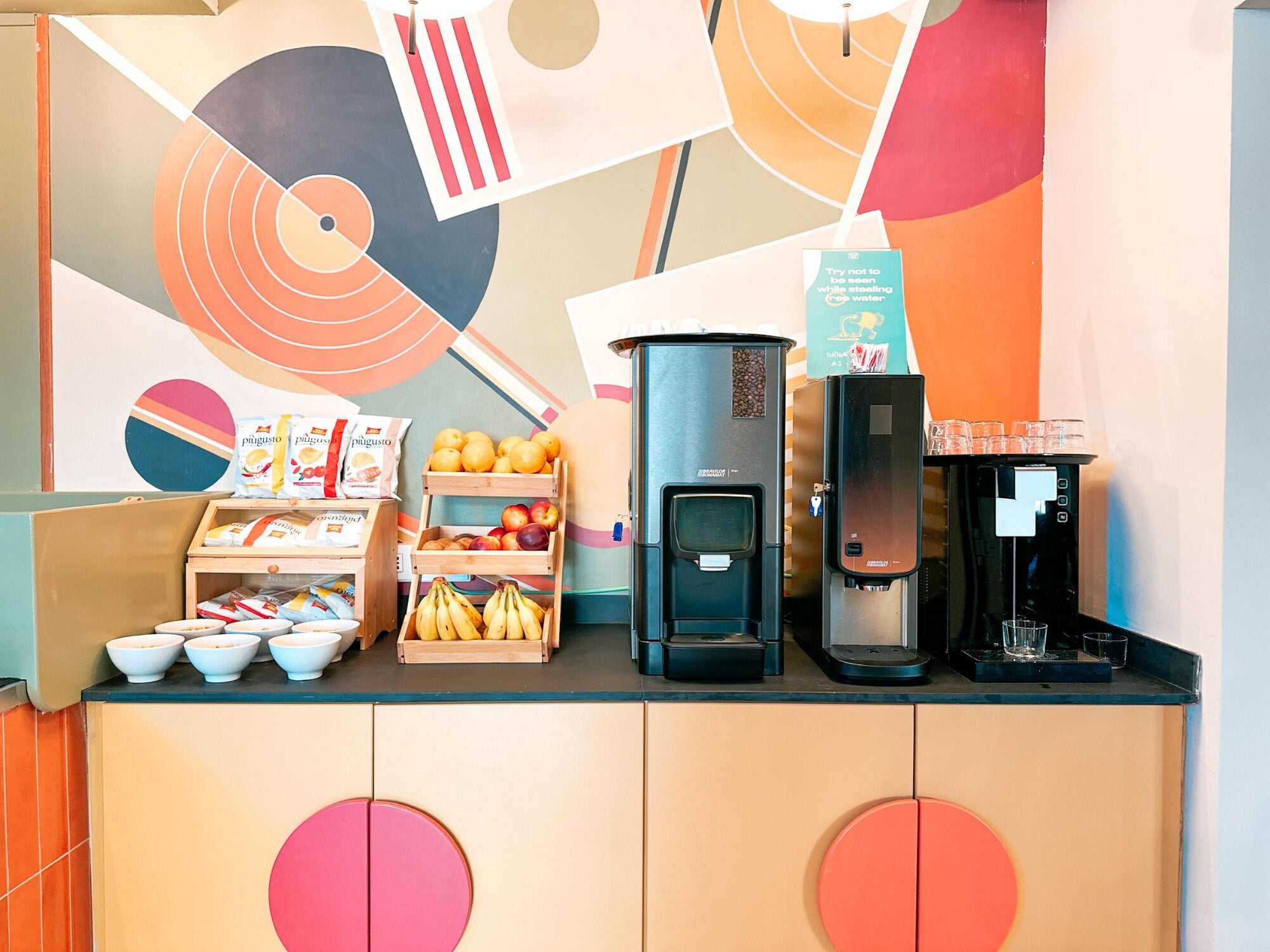 Una estación de café con una máquina de café, dispensador de agua y una exhibición de frutas.