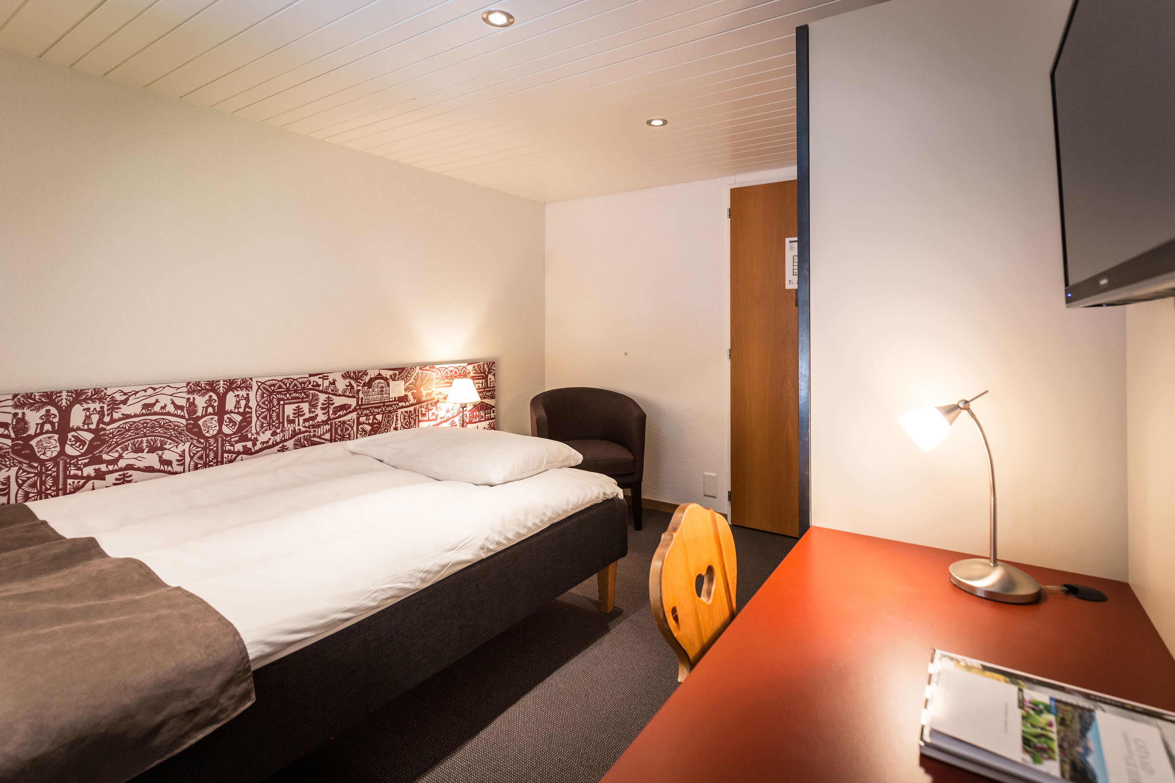 Ein Hotelzimmer mit einem Bett, einem Sessel, einem Schreibtisch mit Lampe und einem an der Wand befestigten Fernseher.