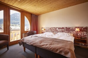Ein Hotelzimmer mit einem großen Bett, einem Sessel, einem Nachttisch mit Lampe und Blick auf Berge durch große Fenster.