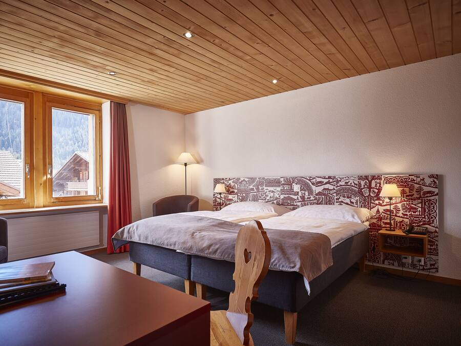 A hotel room with a wooden ceiling, two beds, a desk, a chair, a lamp, and a view of a mountain through large windows.