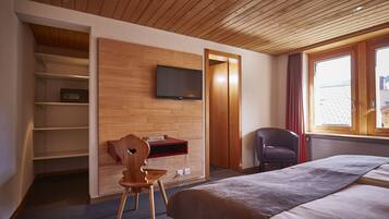 A hotel room with a wooden ceiling, a bed with a grey blanket, a TV mounted on the wall, a wooden desk with a chair, and a window with curtains.