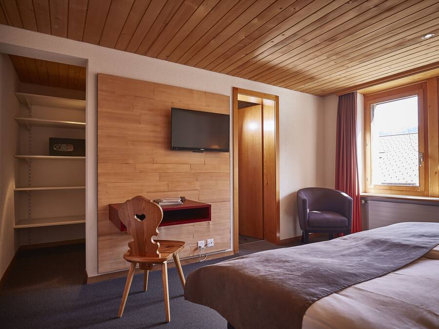 Habitación de hotel con techo de madera, cama con manta gris, televisor montado en la pared, escritorio de madera con una silla y ventana con cortinas.