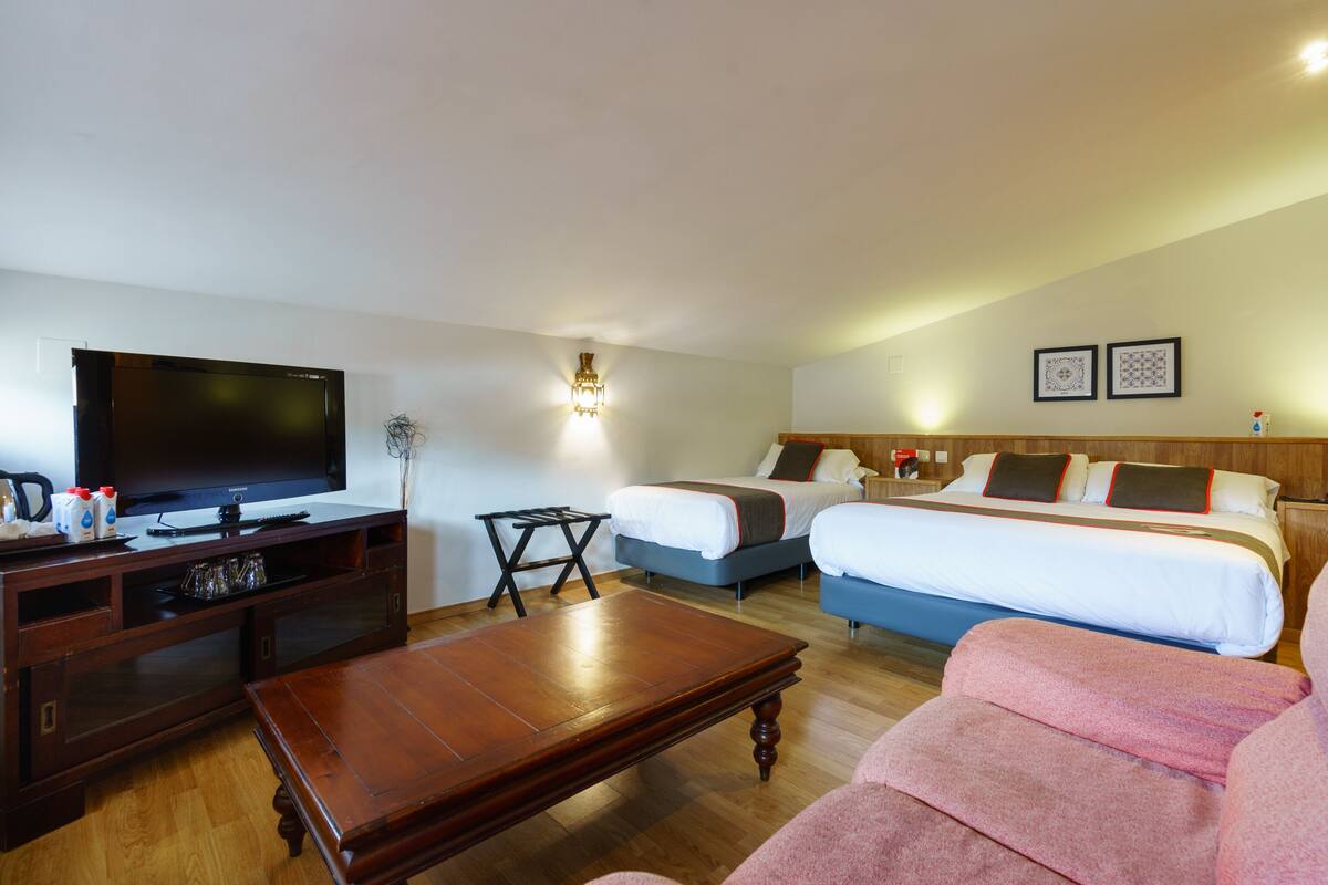 A hotel room with a TV, two beds, a wooden coffee table, and a sofa.
