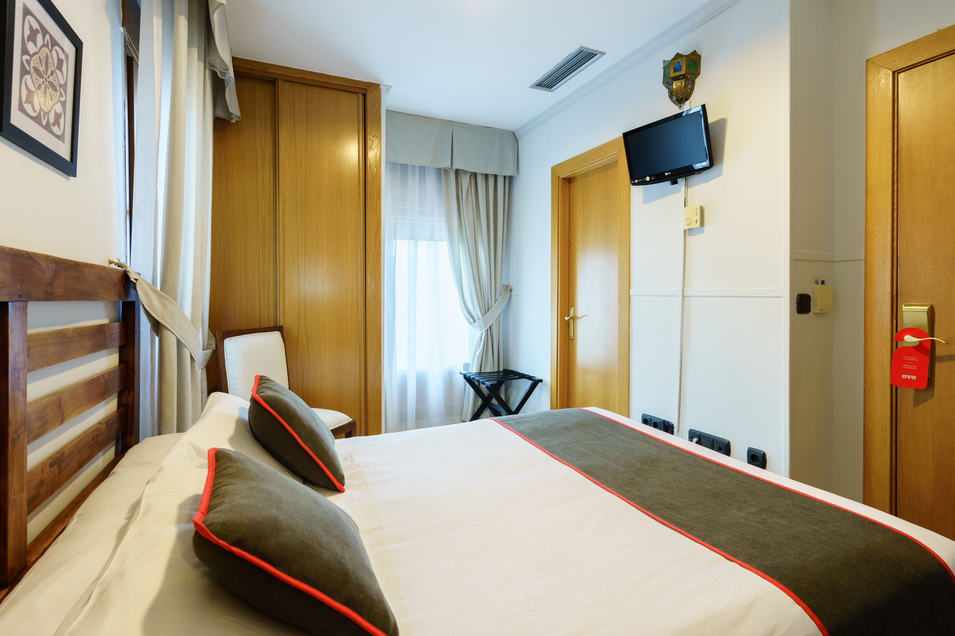 Quarto de hotel com uma cama grande, cabeceira de madeira, uma TV fixada na parede e uma porta com etiqueta vermelha.