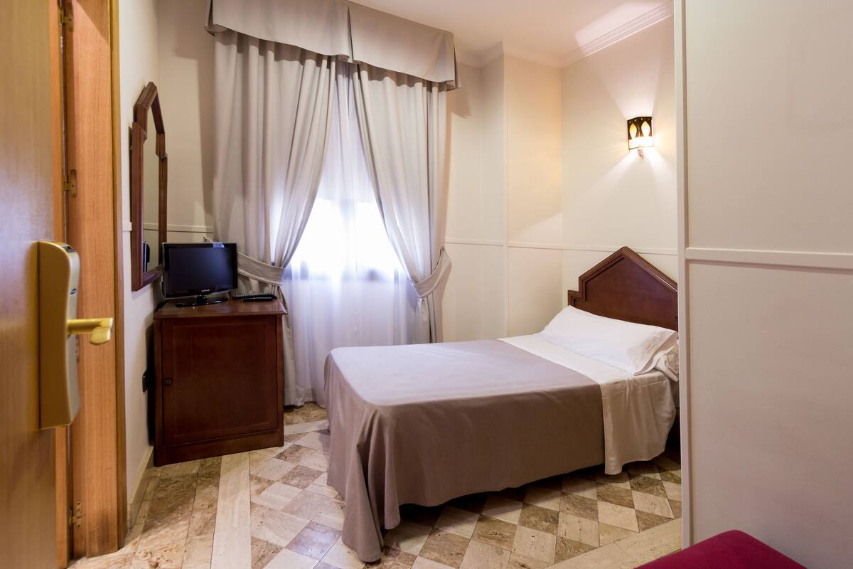 A bedroom with a bed, a nightstand, a television, a mirror, and a window with curtains.