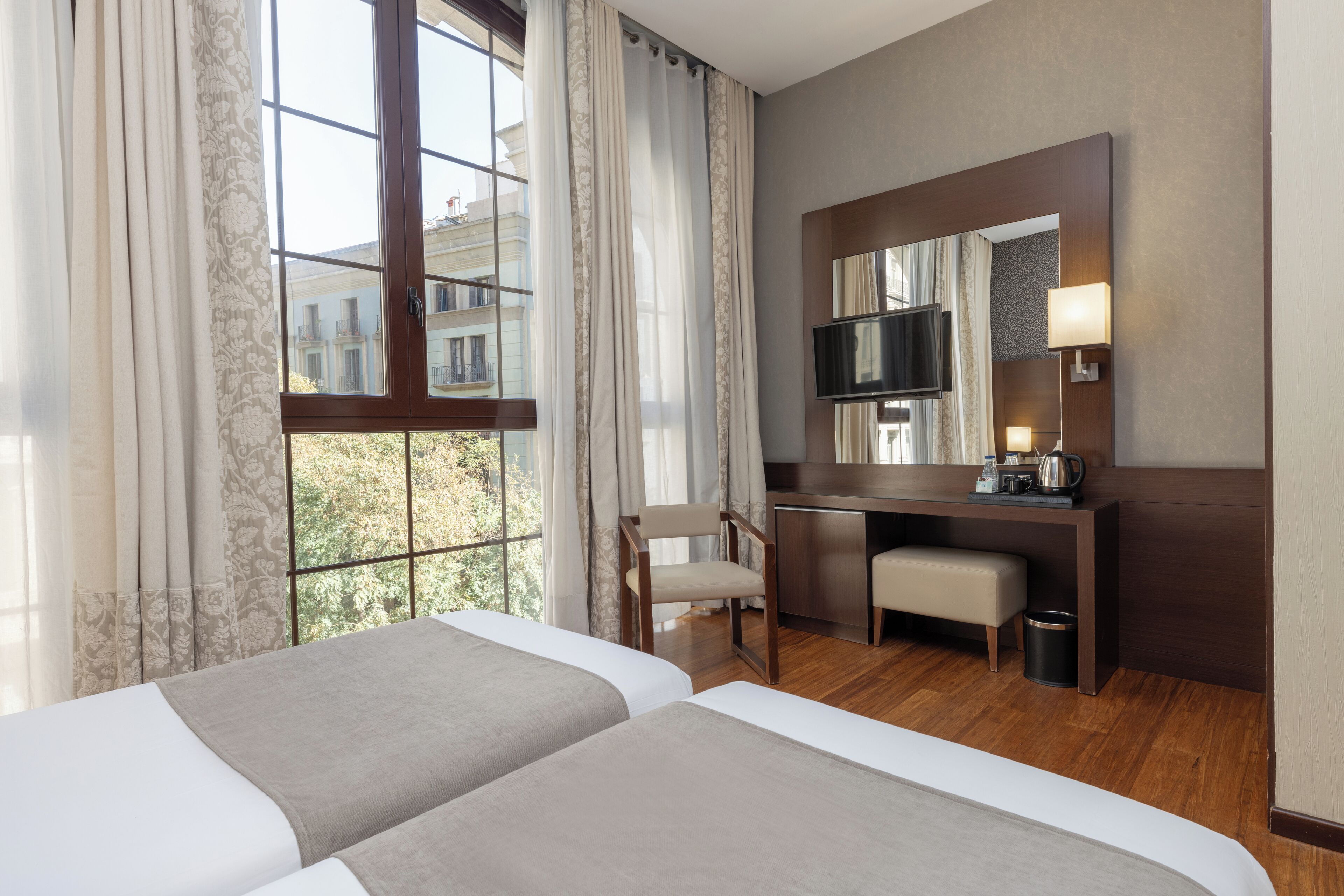 A hotel room with two beds, a desk, a chair, a TV, and a large window with curtains.