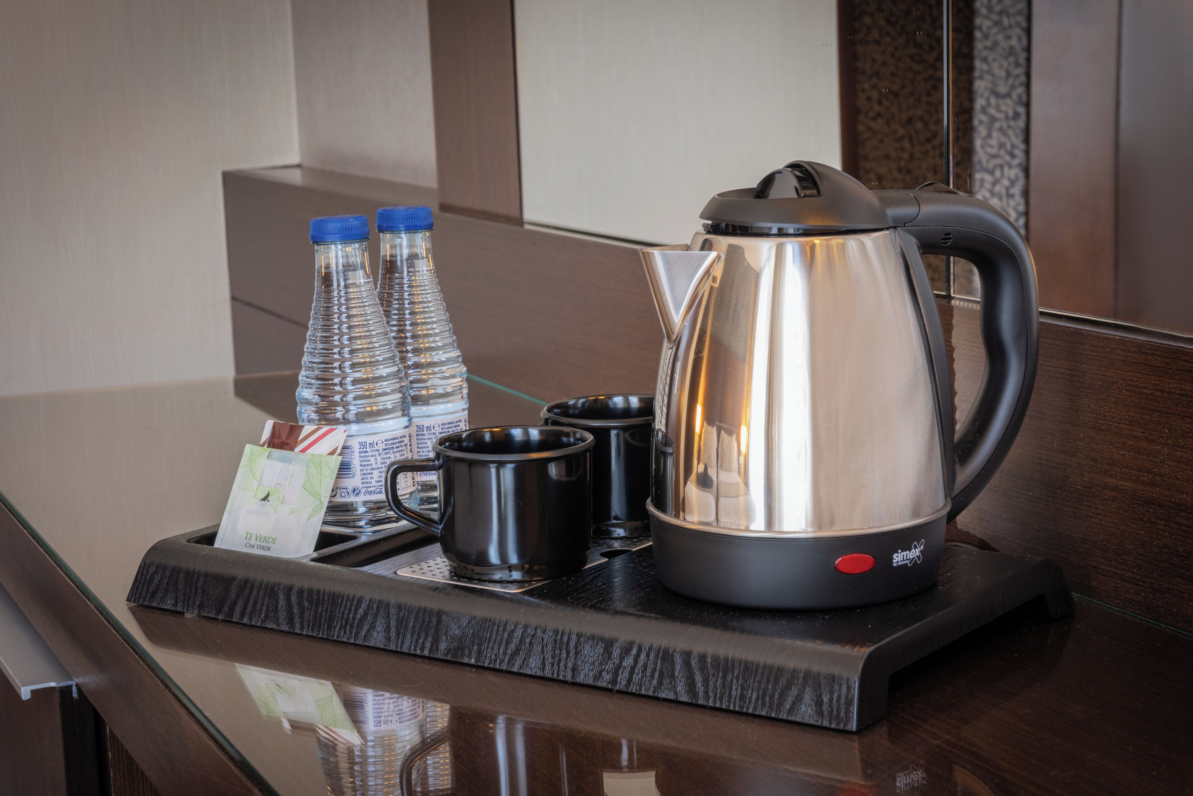 A tray with a stainless steel electric kettle, two black mugs, and two water bottles on a reflective surface.