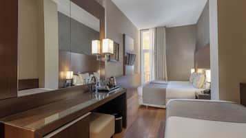 A hotel room with a wooden desk, two beds, a large mirror, a TV, and a window with curtains.