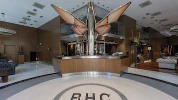 Um hall de hotel moderno com um balcão de recepção central, um grande lustre e um emblema circular com "BHC" no chão.