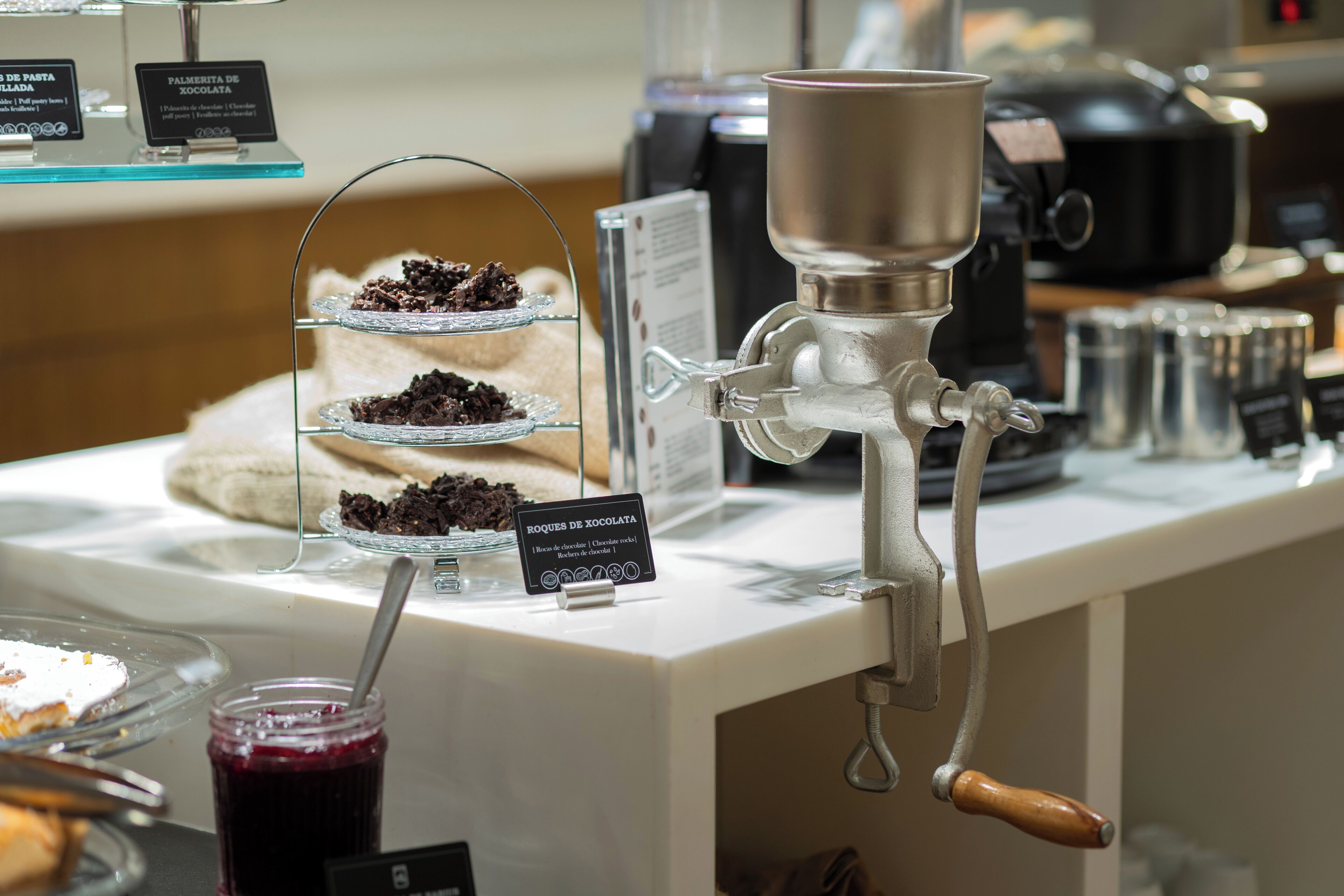 A coffee machine with a metal spout and a wooden handle, a glass display case with layered desserts, and a sign indicating a chocolate dessert.