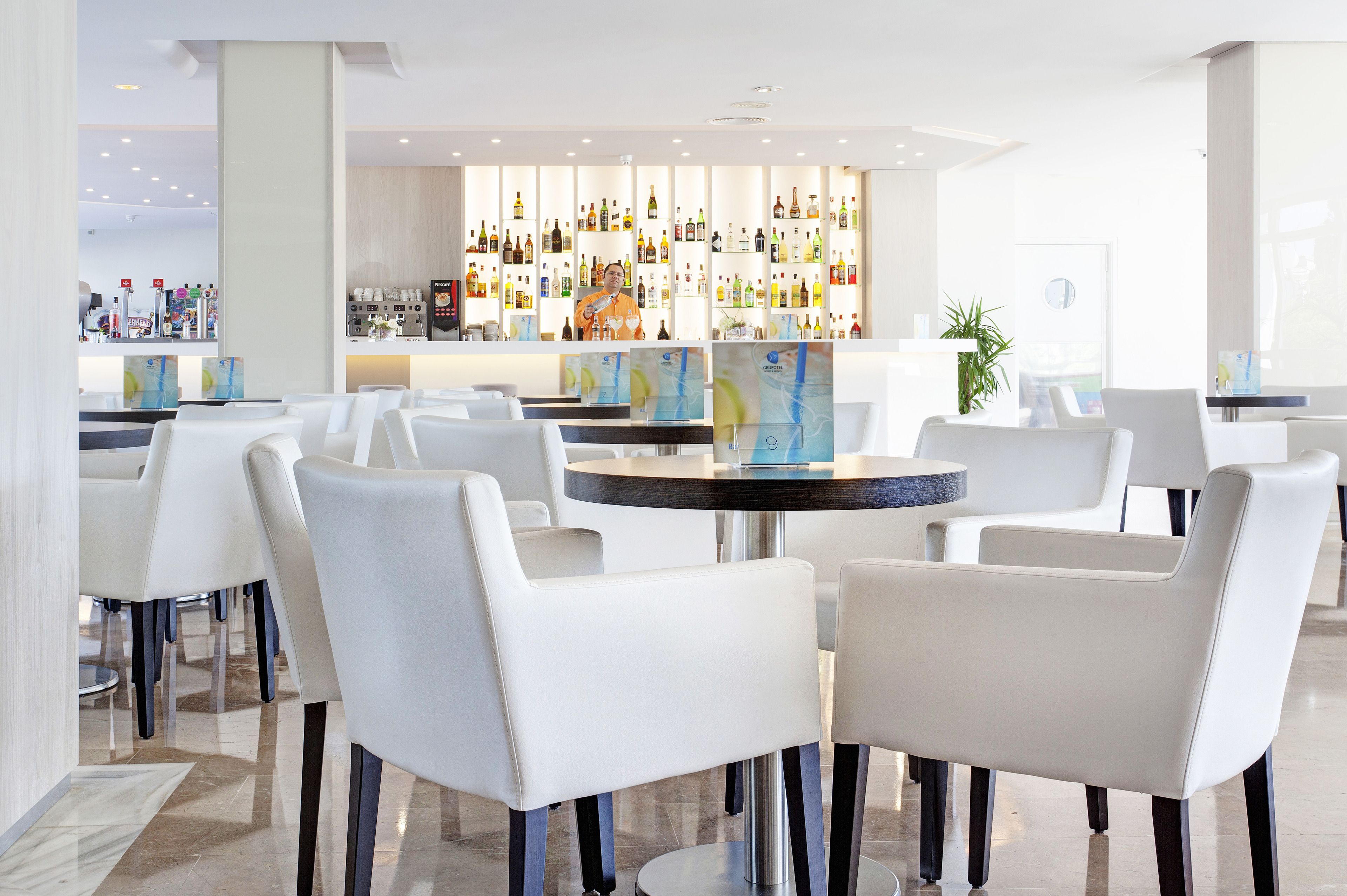 Eine moderne Hotelhalle mit weißen Ledersesseln, einem runden Tisch und einer Bar mit verschiedenen Flaschen und einem großen Gemälde.