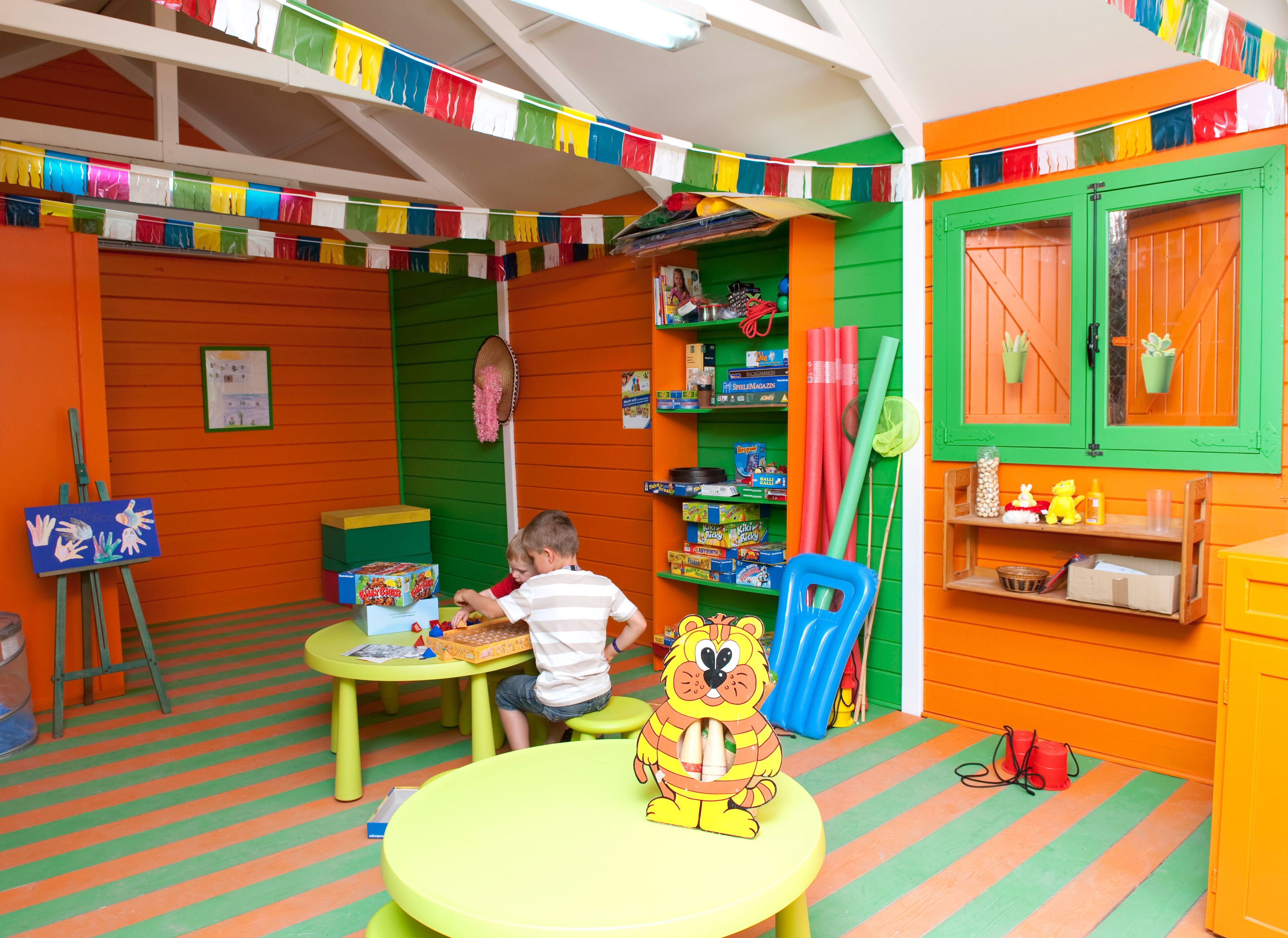 A brightly colored playroom with a child playing at a table, shelves with toys, and a small easel with artwork.