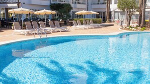 Une piscine d’un bleu éclatant, bordée de chaises longues et d’ombrelles, entourée d’une terrasse carrelée et d’un bâtiment à plusieurs étages.