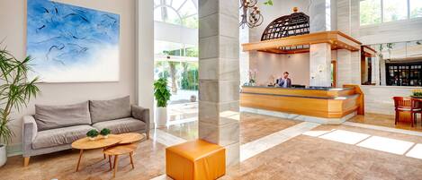 A spacious lobby with a reception desk, a grey sofa, a coffee table, and a large window offering a view of the outdoors.