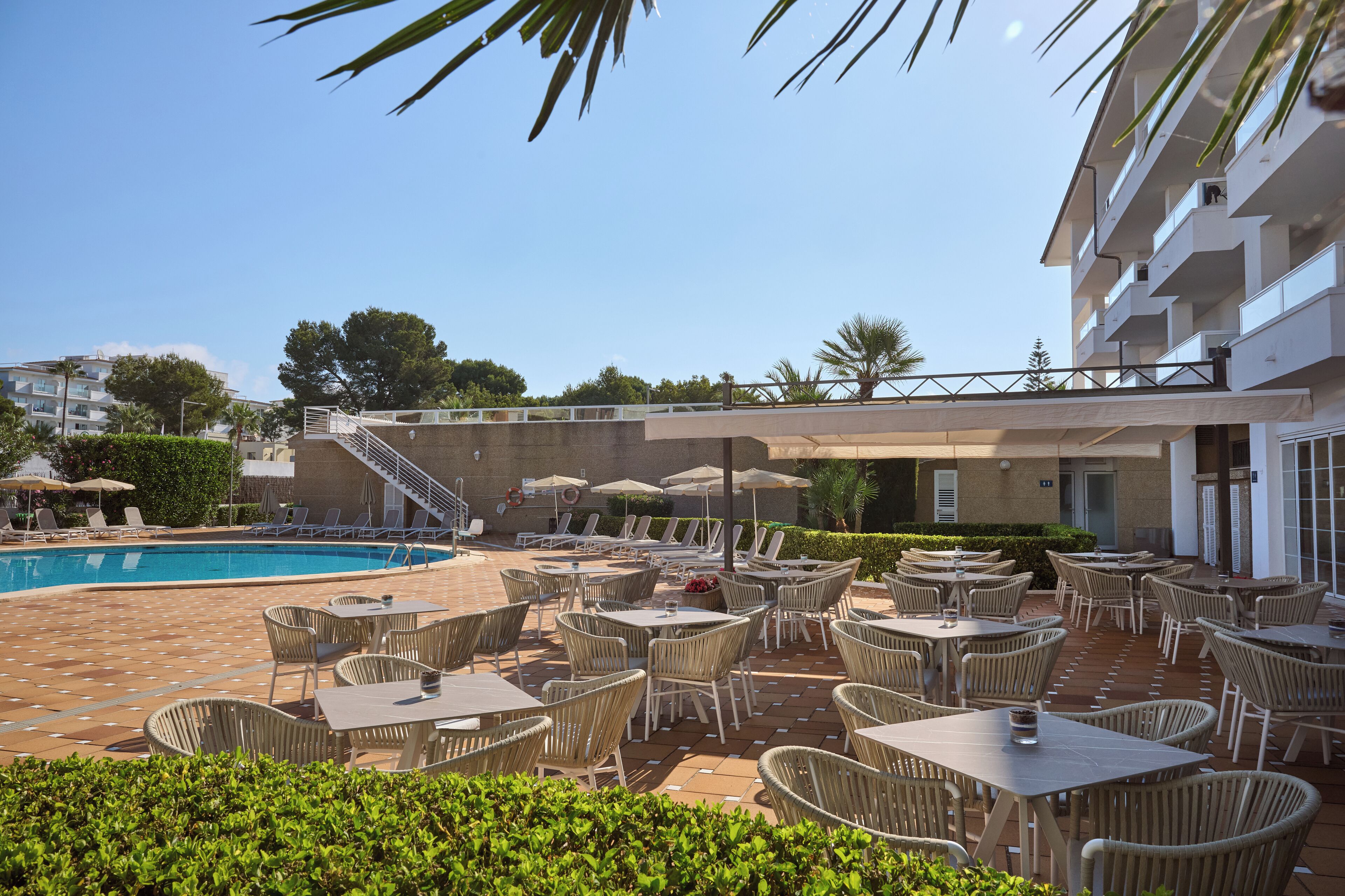 Un área junto a la piscina con sillones y mesas, una escalera que lleva a un nivel superior, y un edificio con balcones.