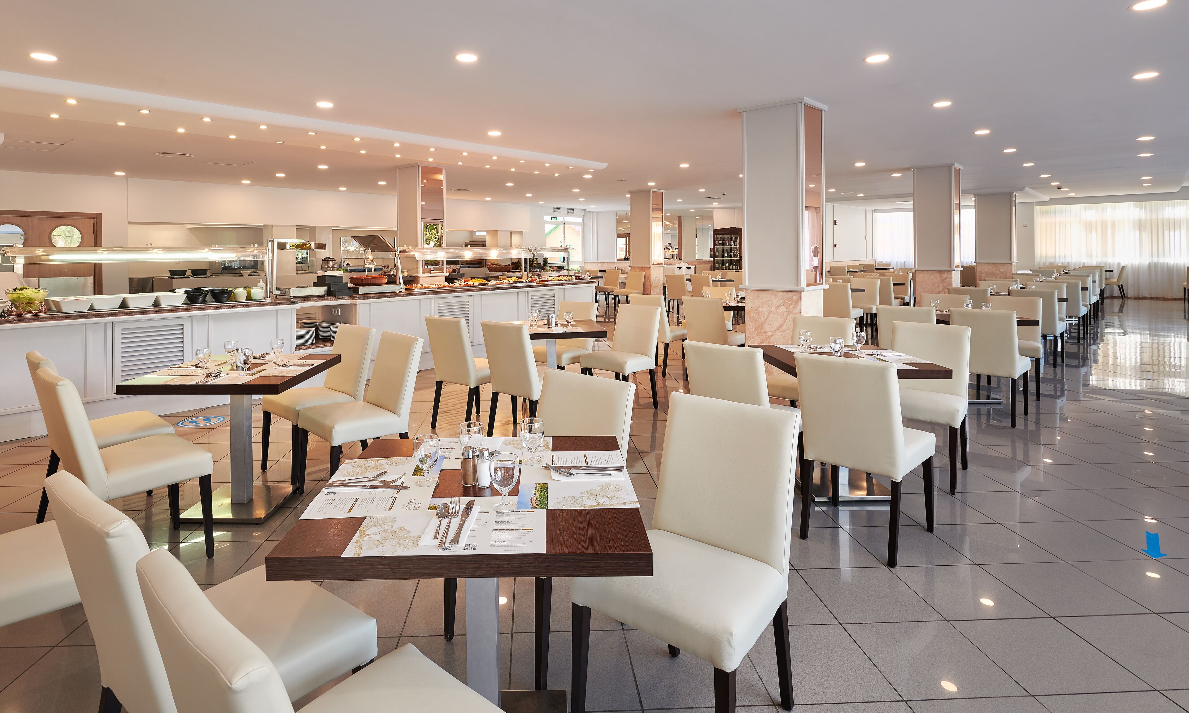 A spacious dining area with multiple tables and chairs, a buffet with various food items, and a modern kitchen in the background.