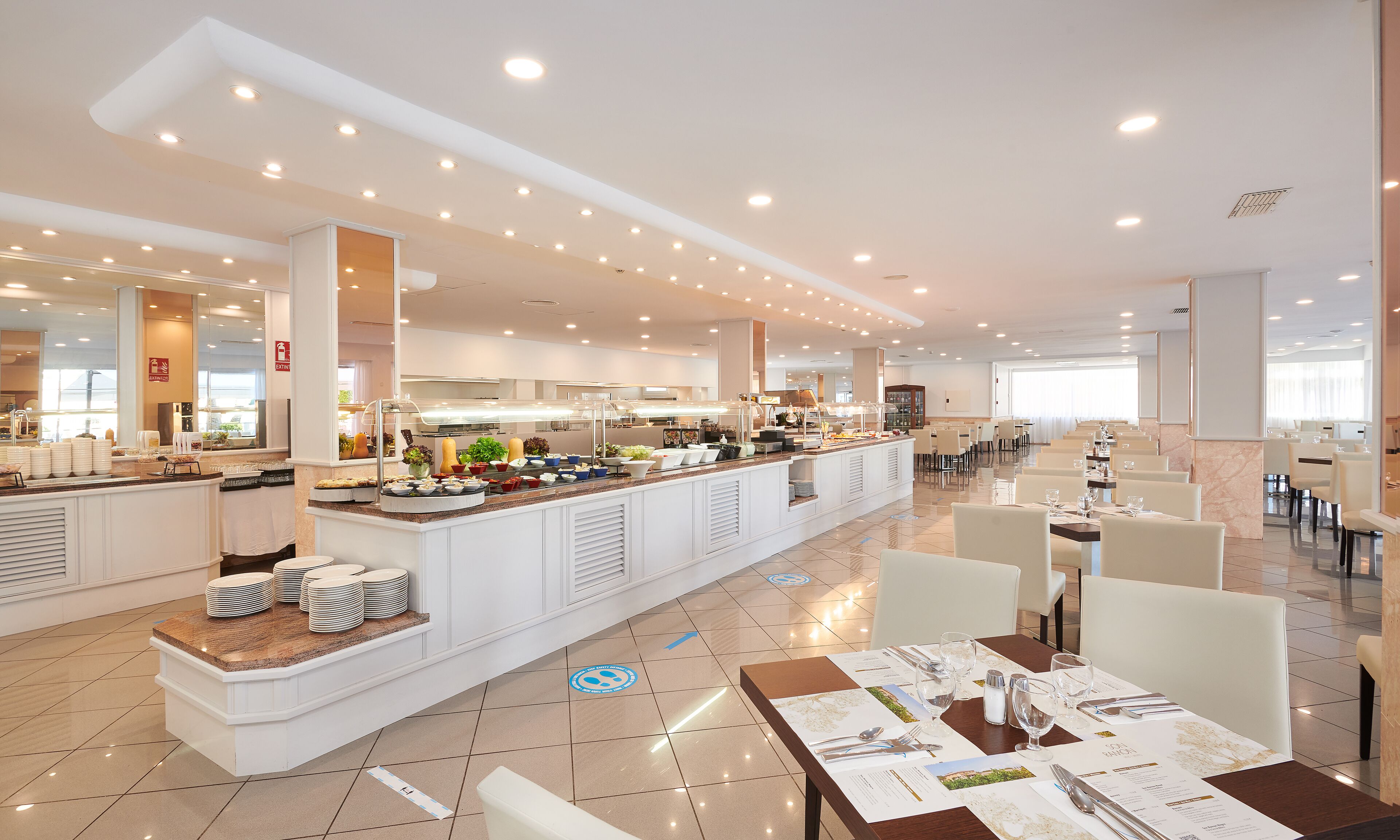 A spacious hotel dining area with a buffet, multiple seating arrangements, and a modern kitchen in the background.