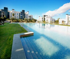 A modern swimming pool with a clear blue water surface, surrounded by a well-maintained grassy area and a row of contemporary buildings in the background. - El Plantio Golf Resort (Alicante)