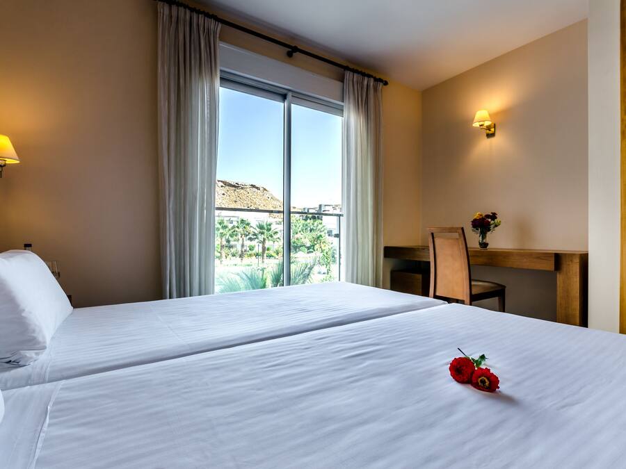 A hotel room with two beds, a desk, a chair, a lamp, and a window with curtains.