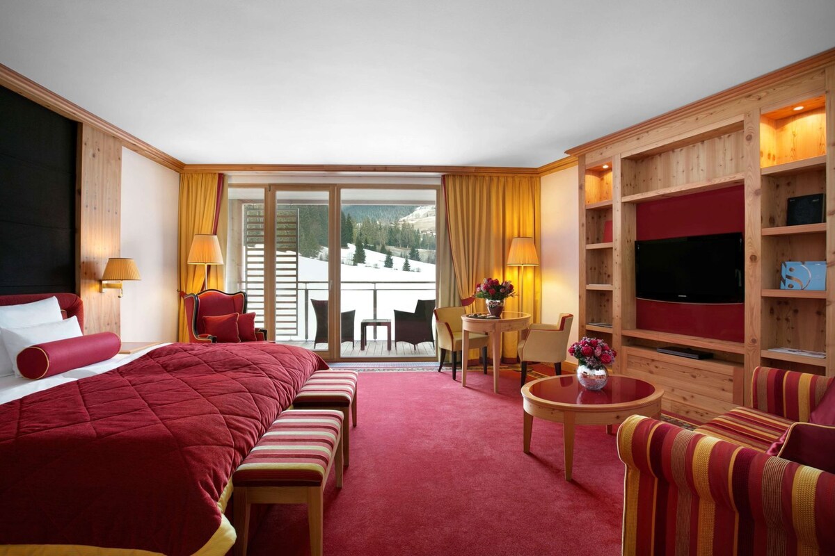 Habitación de hotel con una cama grande, un escritorio, un televisor y un balcón con vistas a las montañas.