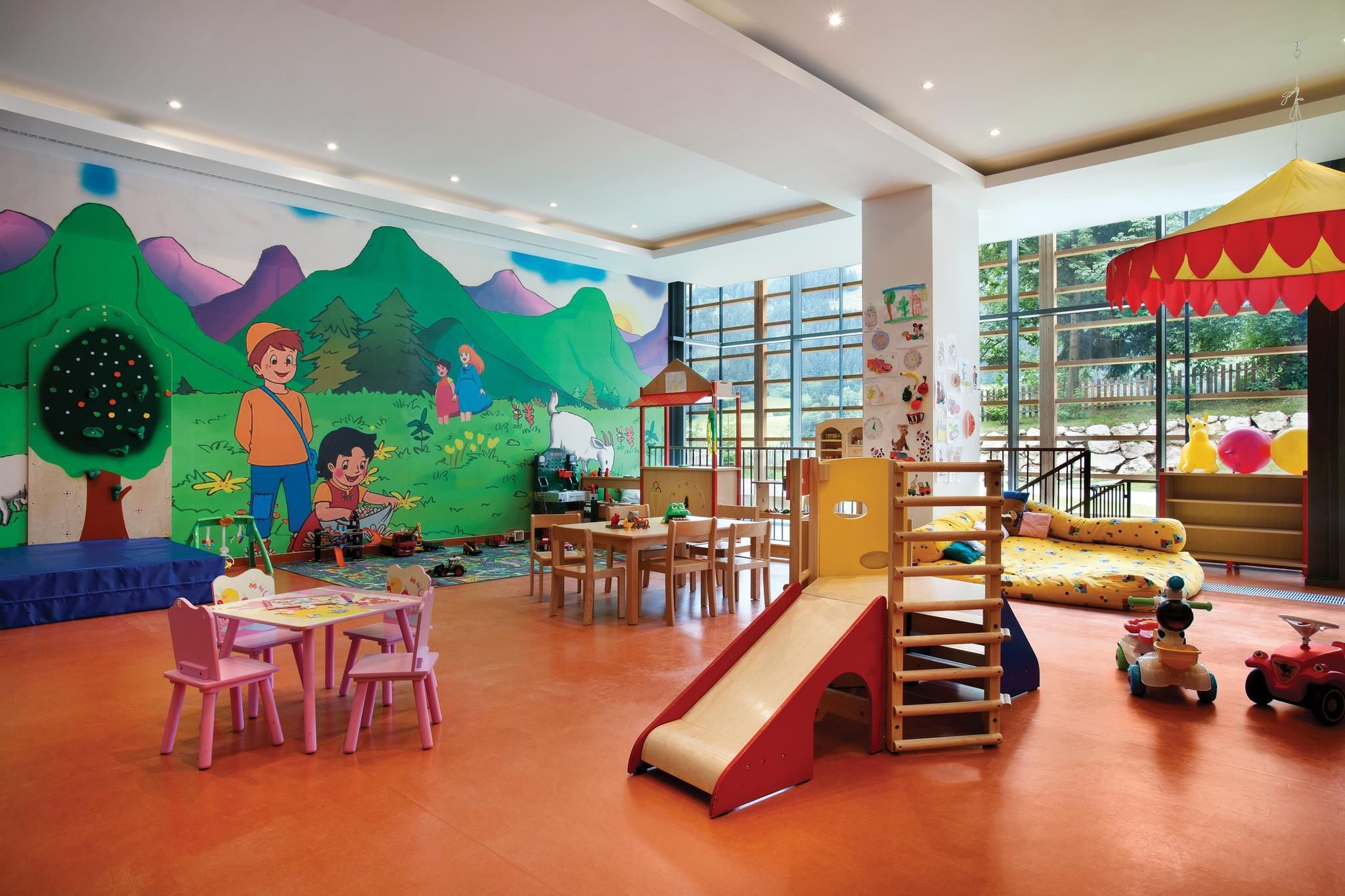 Un área de juegos infantiles con un tobogán, mesas, sillas y un mural en la pared.