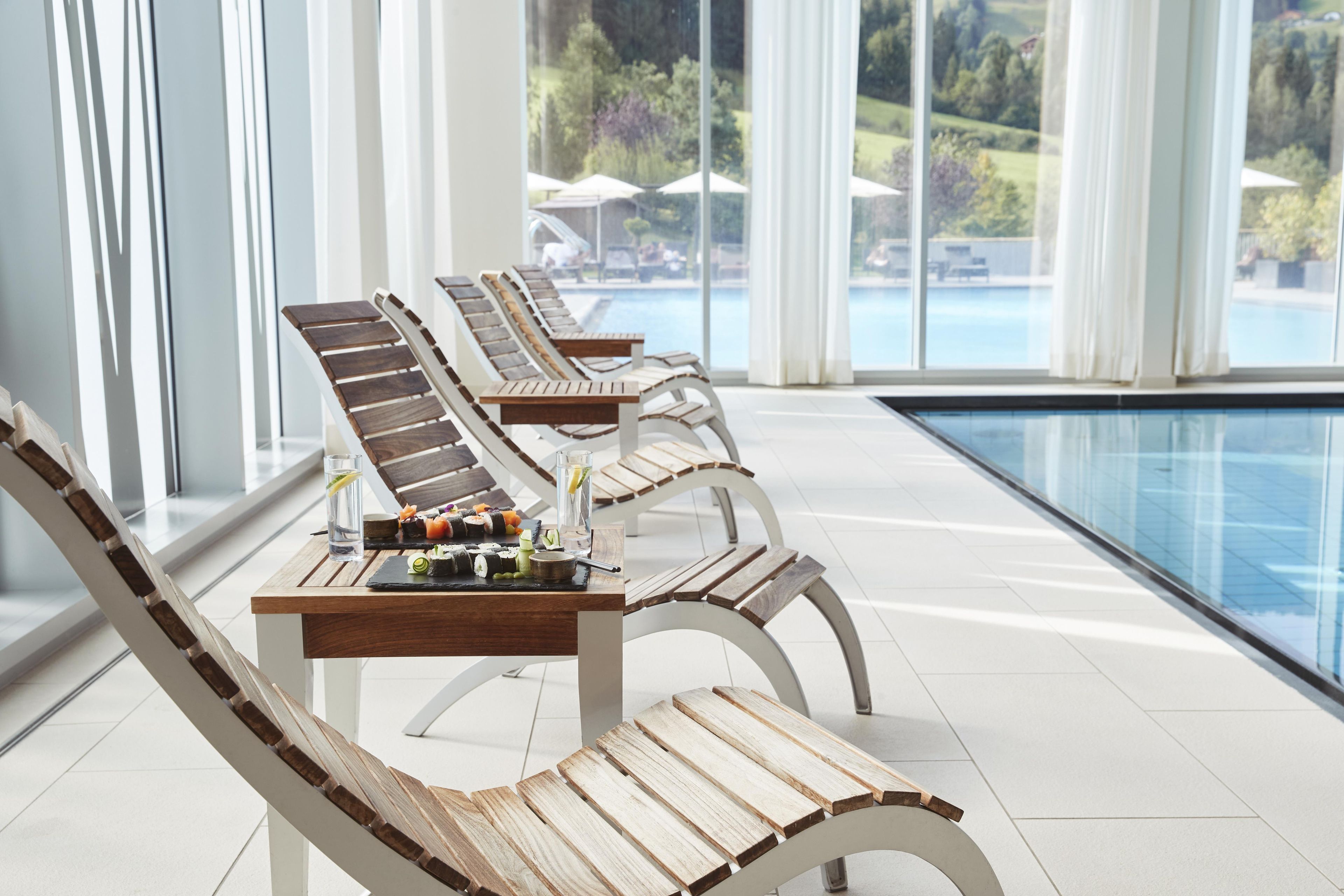 A row of wooden lounge chairs by a poolside with a clear view of the pool and greenery outside.