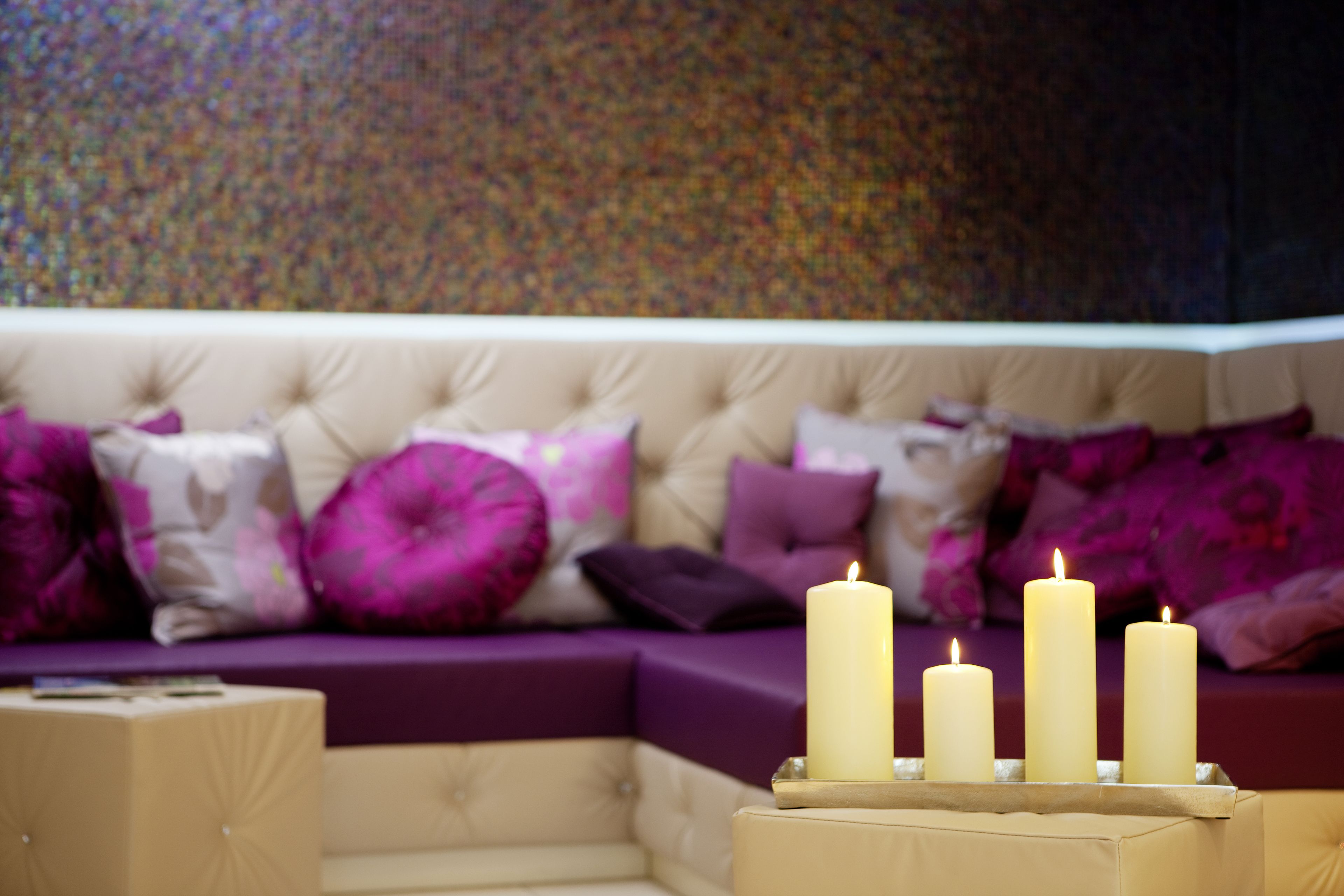 A cozy living room with a beige sofa adorned with purple and pink cushions, and three lit candles on a wooden side table.
