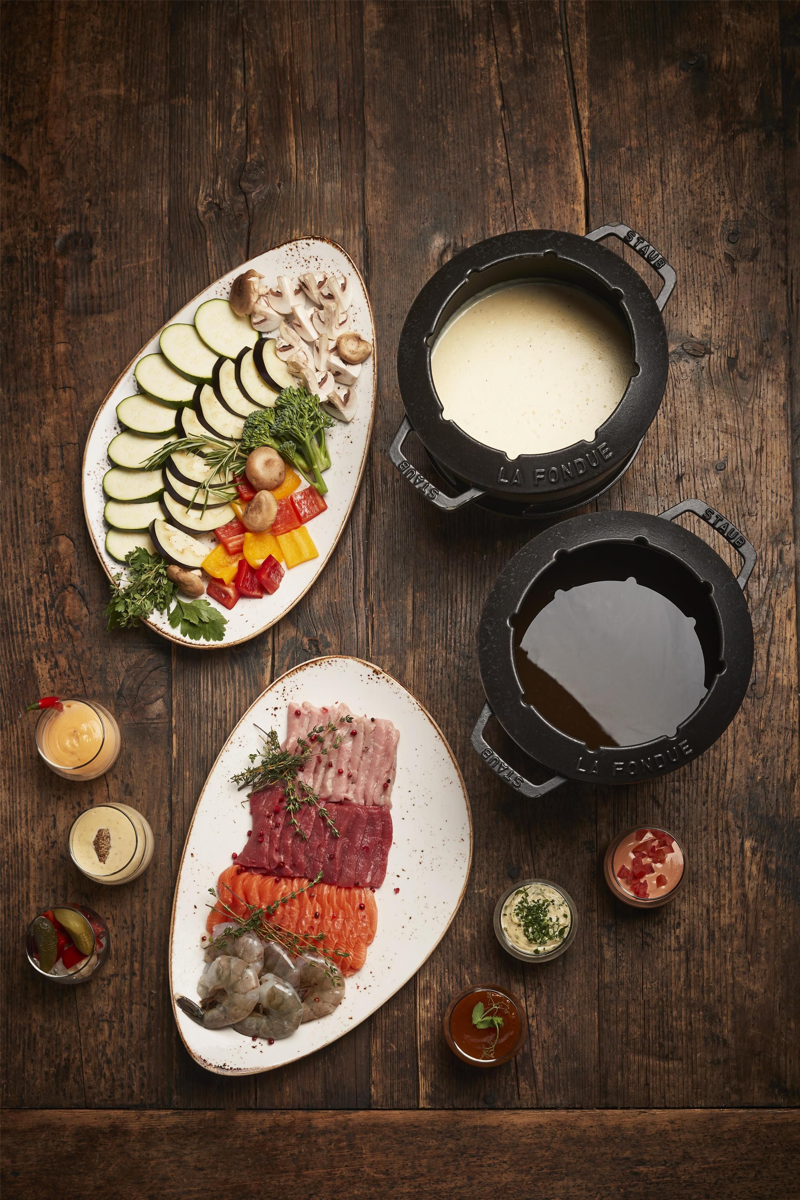 A wooden table with a variety of food items, including sliced vegetables, meats, and seafood, accompanied by two black pots with sauces.